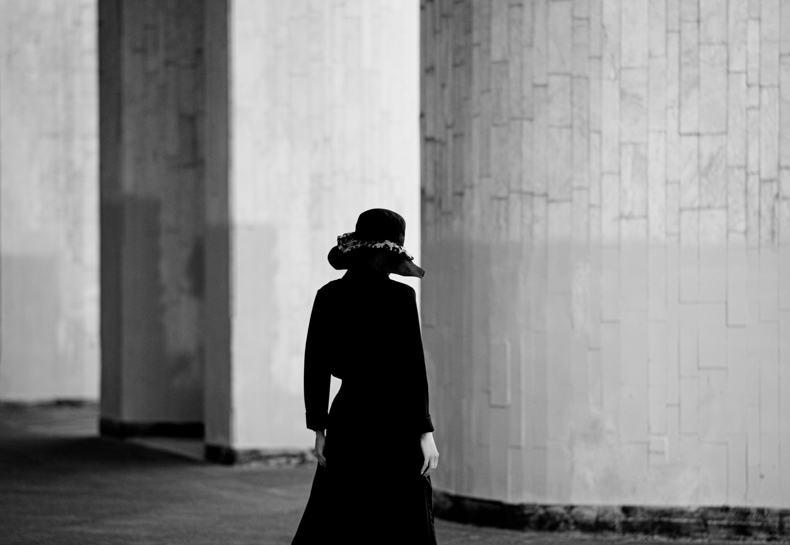Женщина в чёрной шляпе и платье идёт вдоль бетонной стены, чёрно-белая минималистичная художественная фотография