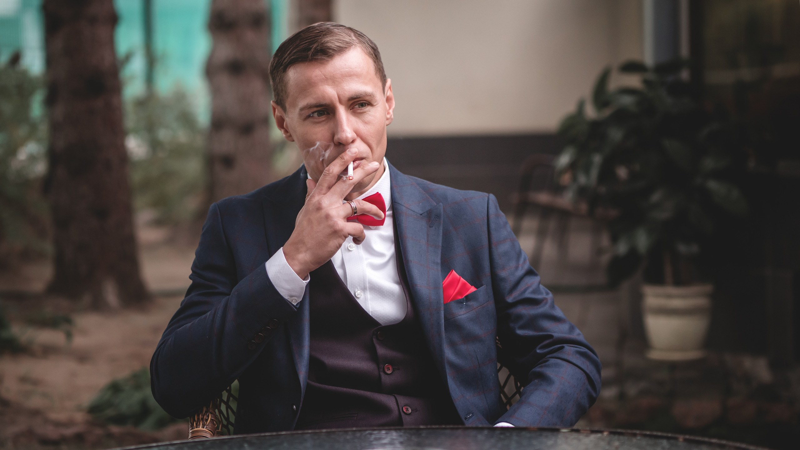 мужчина в строгом синем костюме в полоску и в красной бабочке. на нём белая рубашка и сидит он за столом курит сигарету.