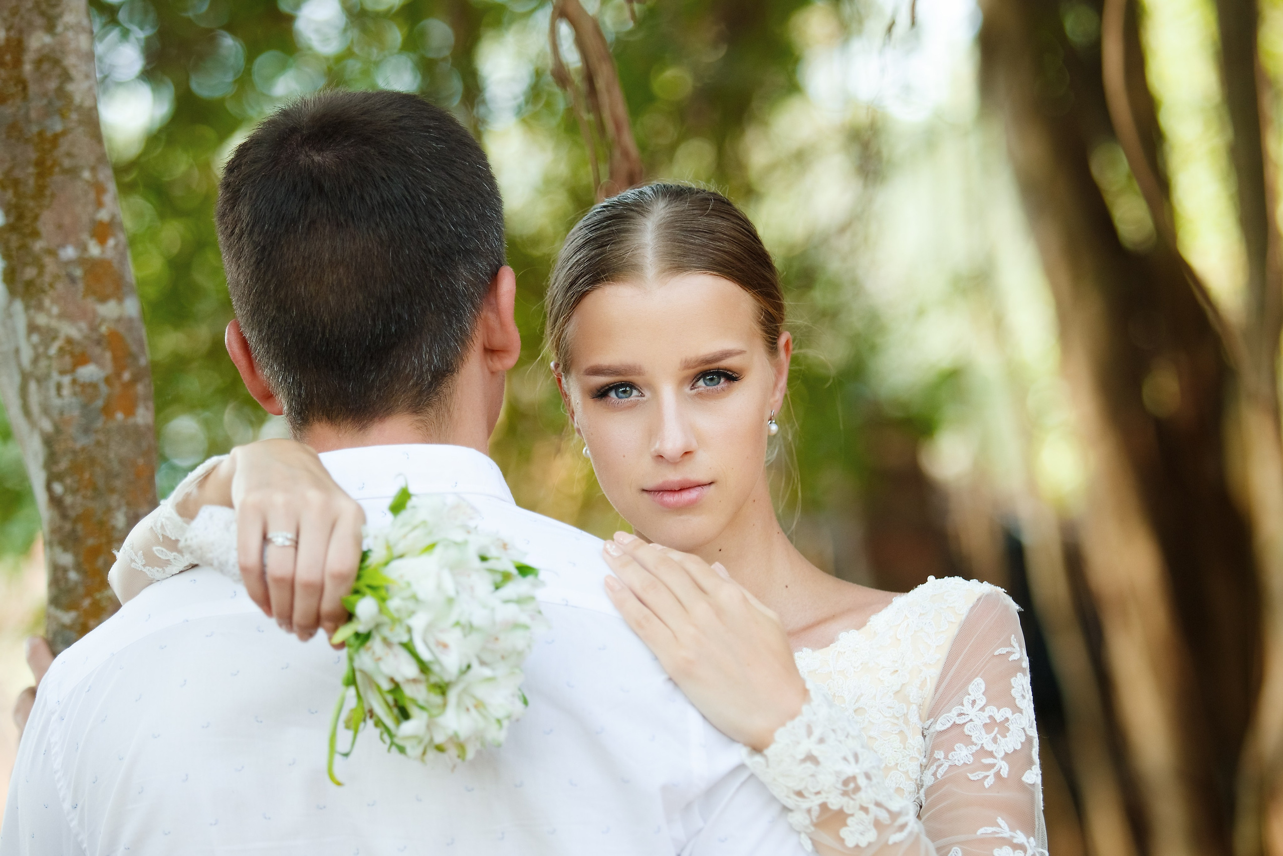 Невеста в кружевном платье обнимает жениха. Теплый взгляд, нежные руки, фон — размытые огни.