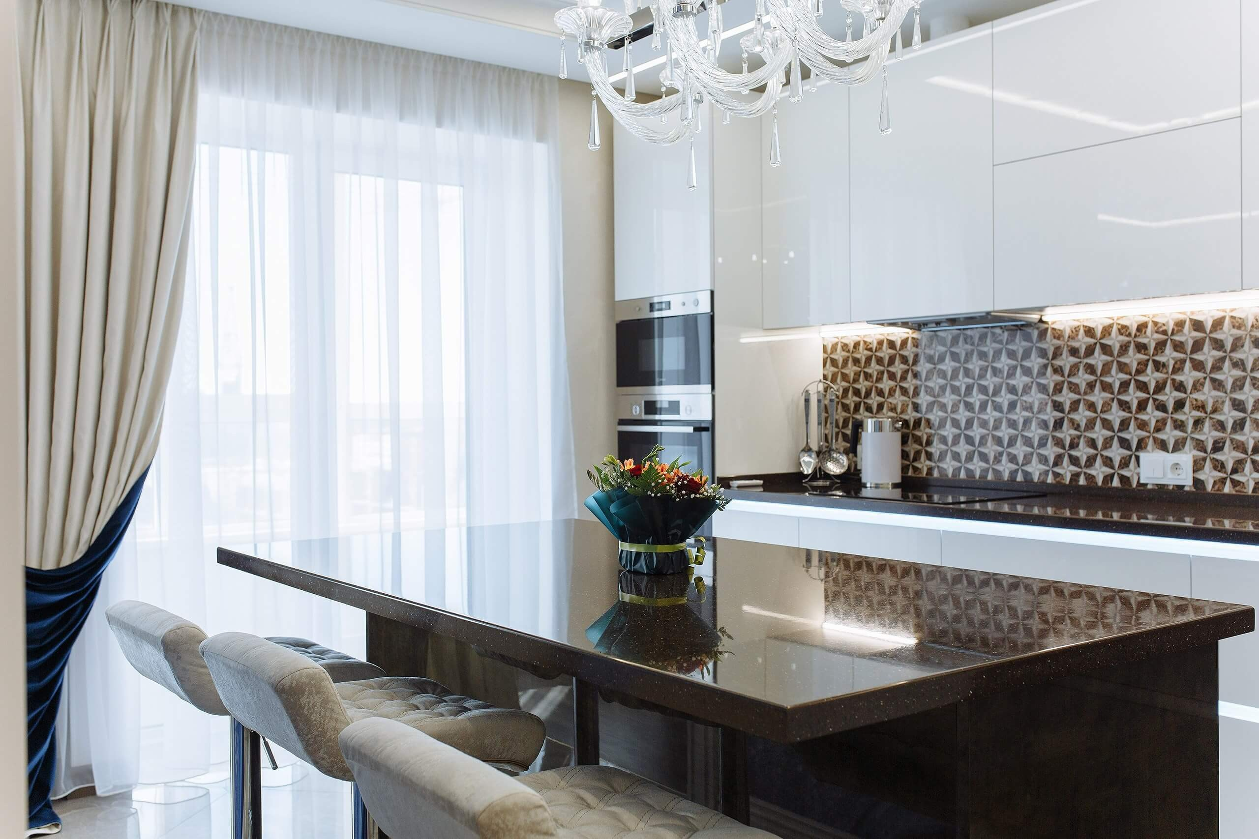 Фотография кухни в стиле современная классика: белый глянцевый гарнитур, остров со столешницей из темного кварца и барные стулья с обивкой капитоне. Акцентный фартук с 3D-узором. Фото для каталога кухонной студии в Пензе.