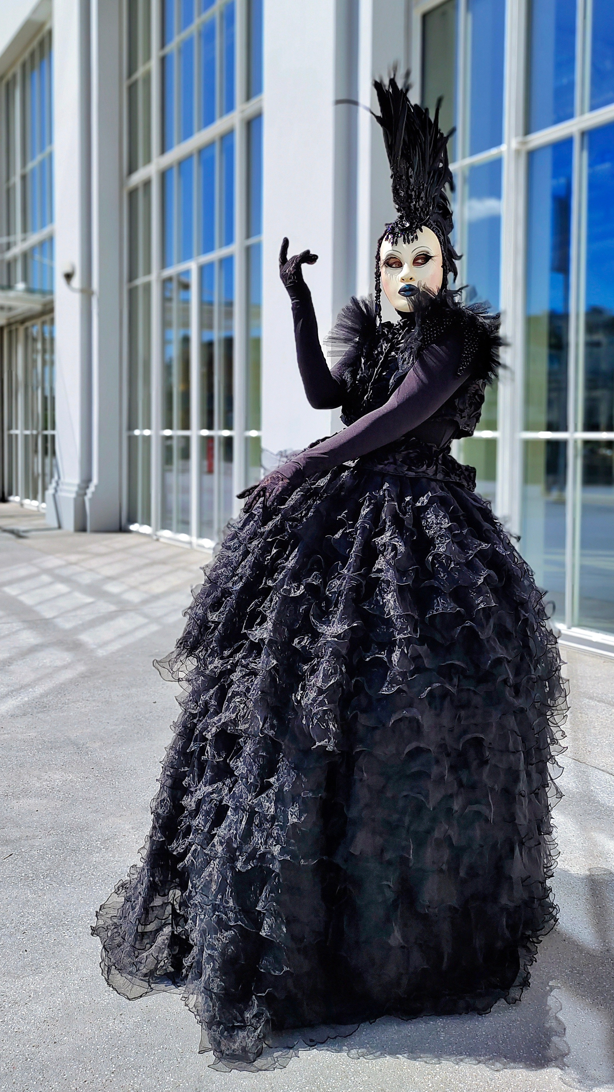 Артист мим в чёрном платье и в маске на лице позирует праздничному фотографу.