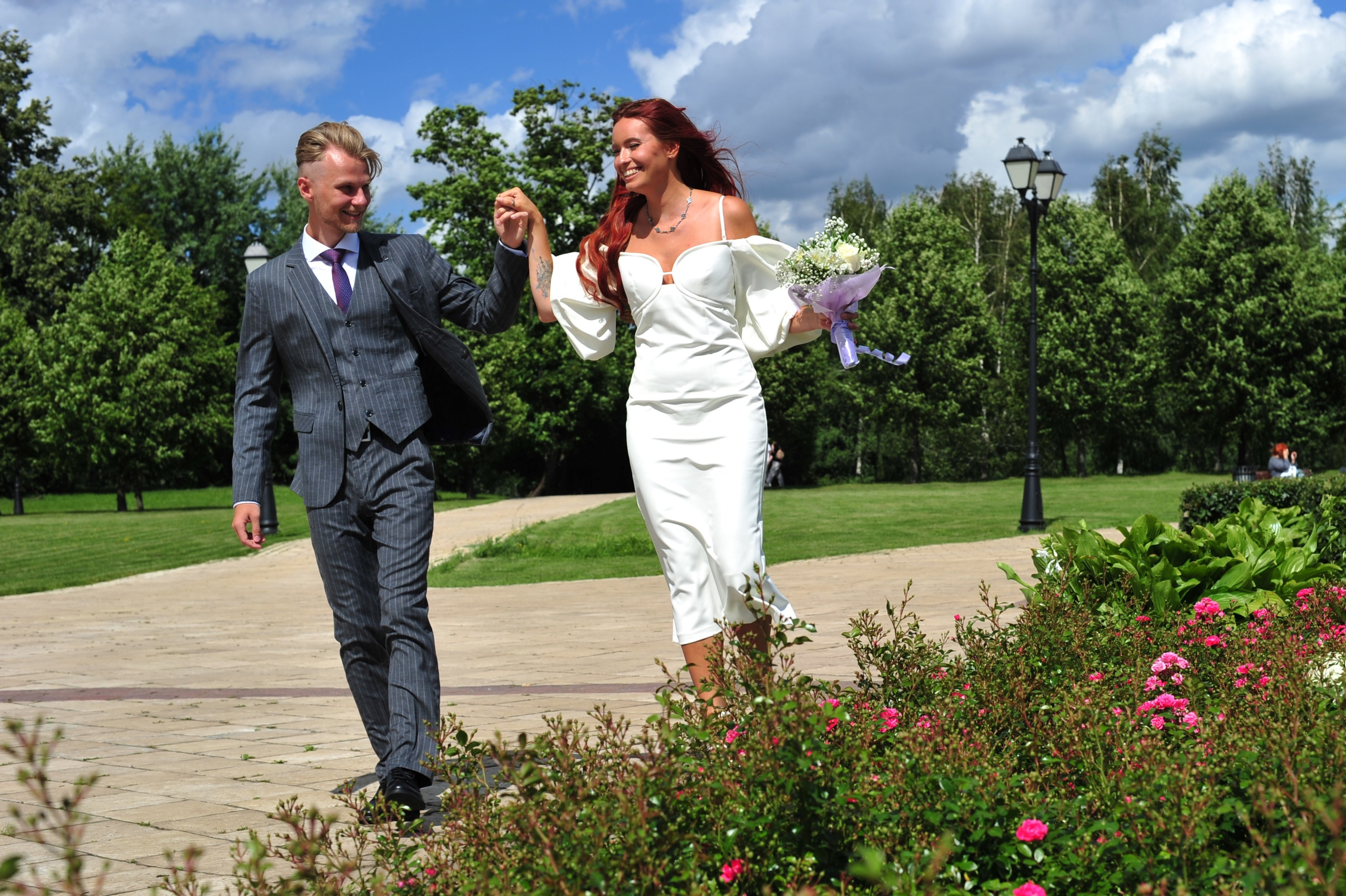 Свадьба в Царицыно летом, жених ведет невесту за руку по бордюру клумбы