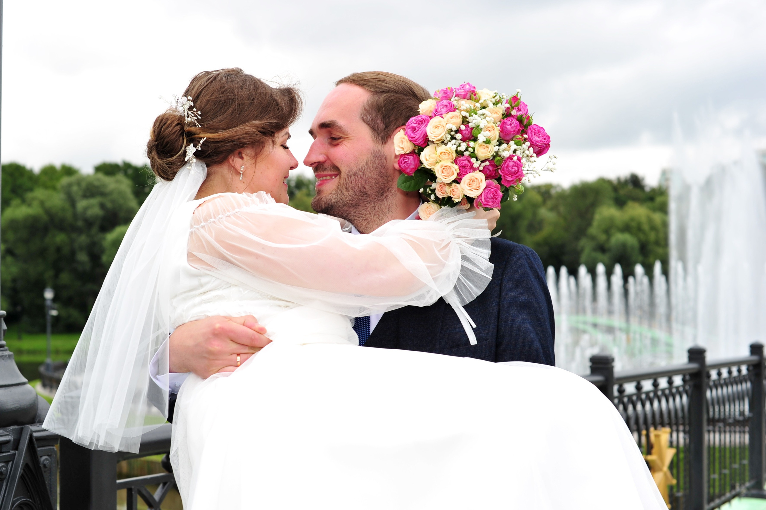 Свадьба в Царицыно пасмурным летним днем, жених держит невесту на руках на железном мосту на фоне фонтанов 
