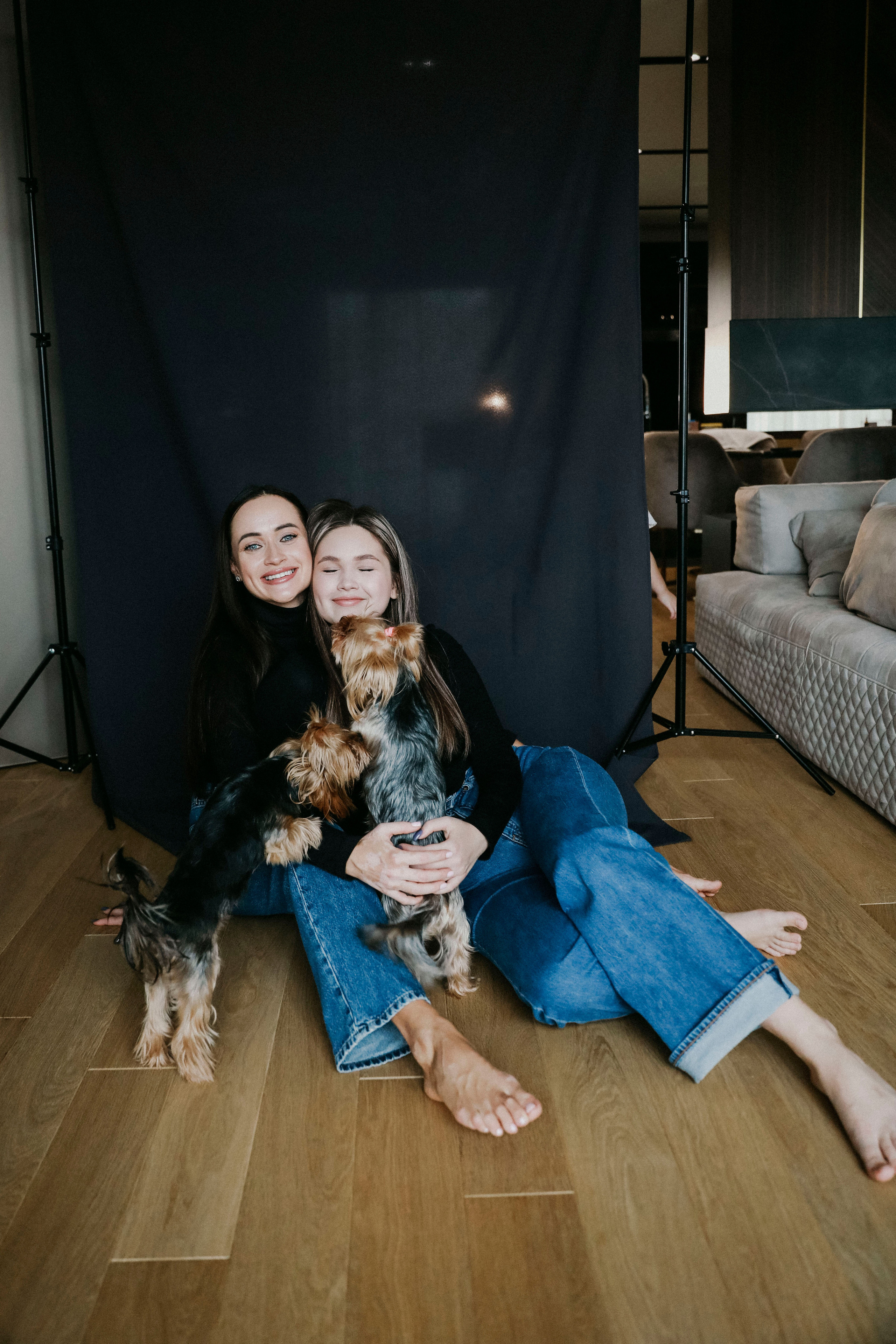 две красивых сестры в джинсах и черных водолазках  с играют с собакой у себя дома