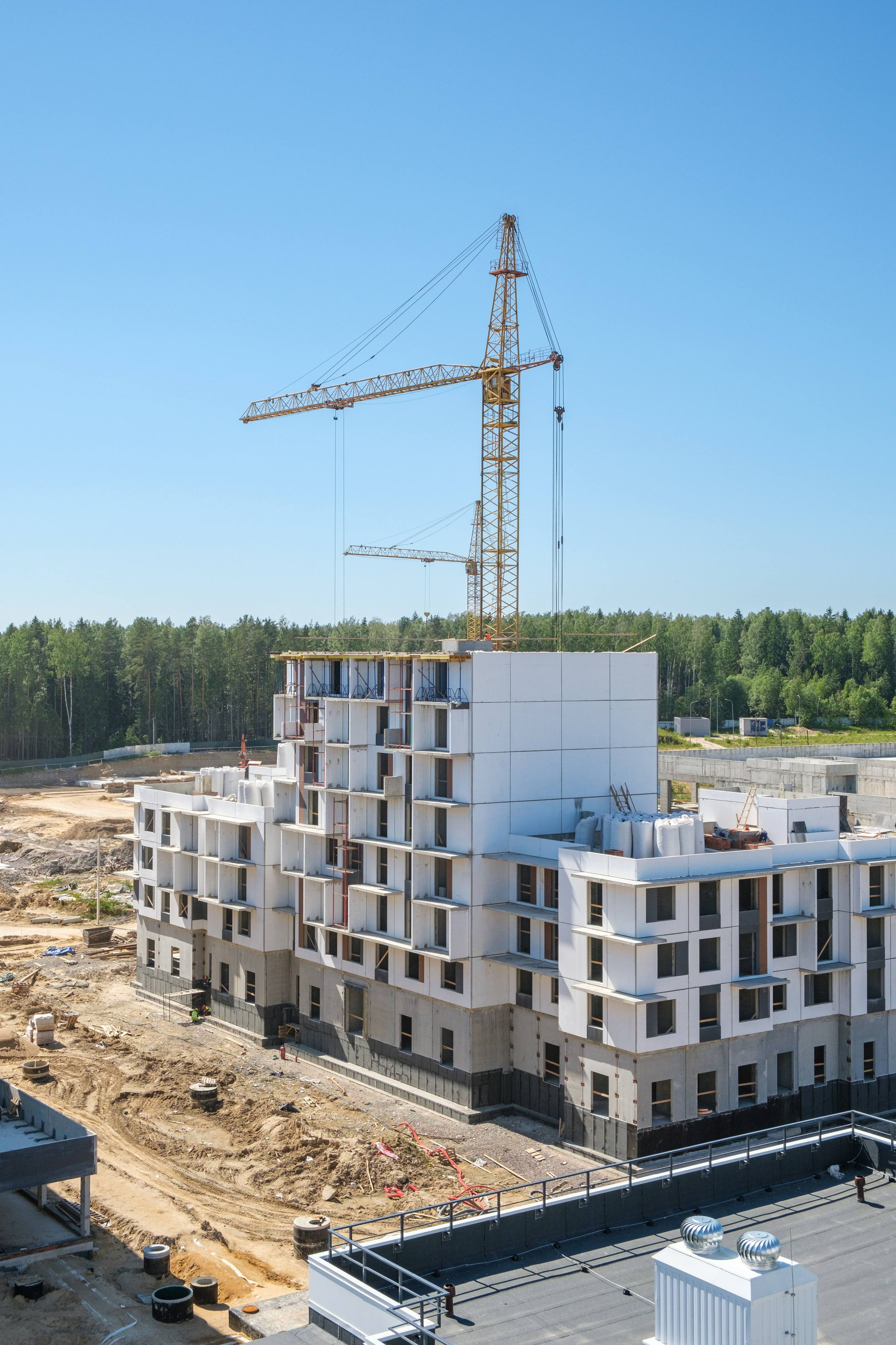 На изображении представлен строящийся многоквартирный жилой комплекс. На переднем плане видны строительные краны и земляные работы, что свидетельствует о том, что строительство еще не завершено.