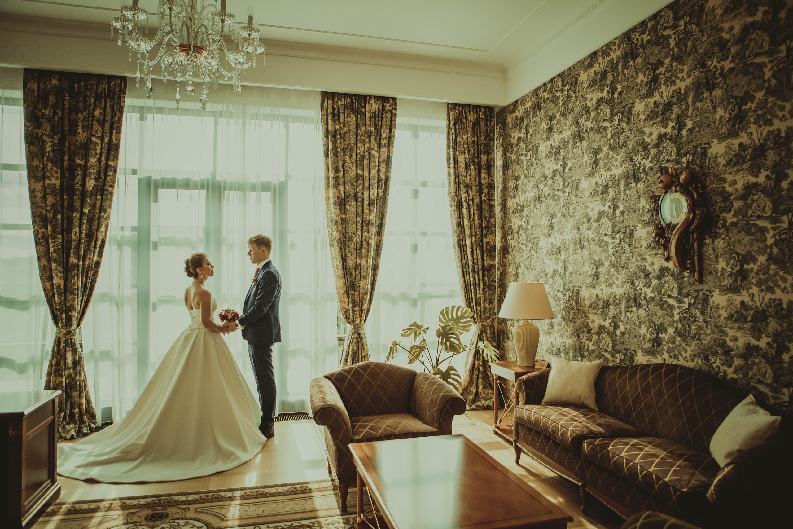 жених держит невесту за руки перед гигантским окном в фешенебельной комнате