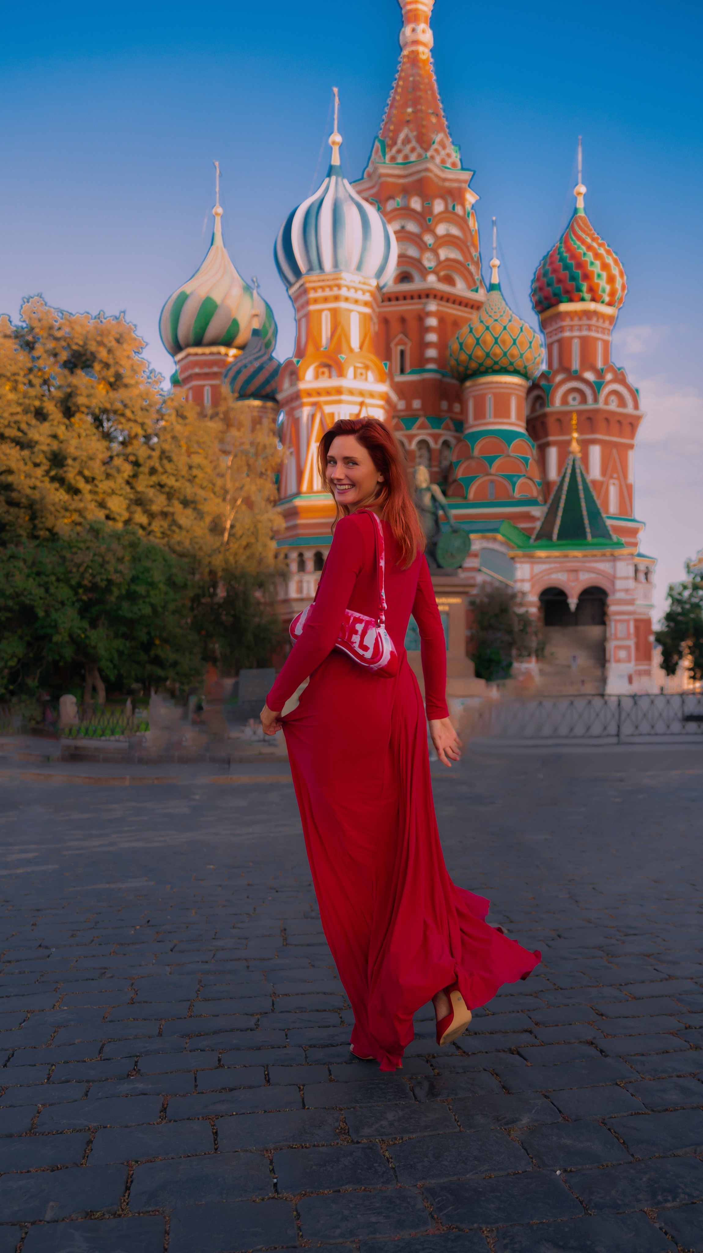 Женщина в длинном струящемся красном платье улыбается через плечо на мощеной площади с собором Василия Блаженного в Москве, Россия, на заднем плане, на закате.
