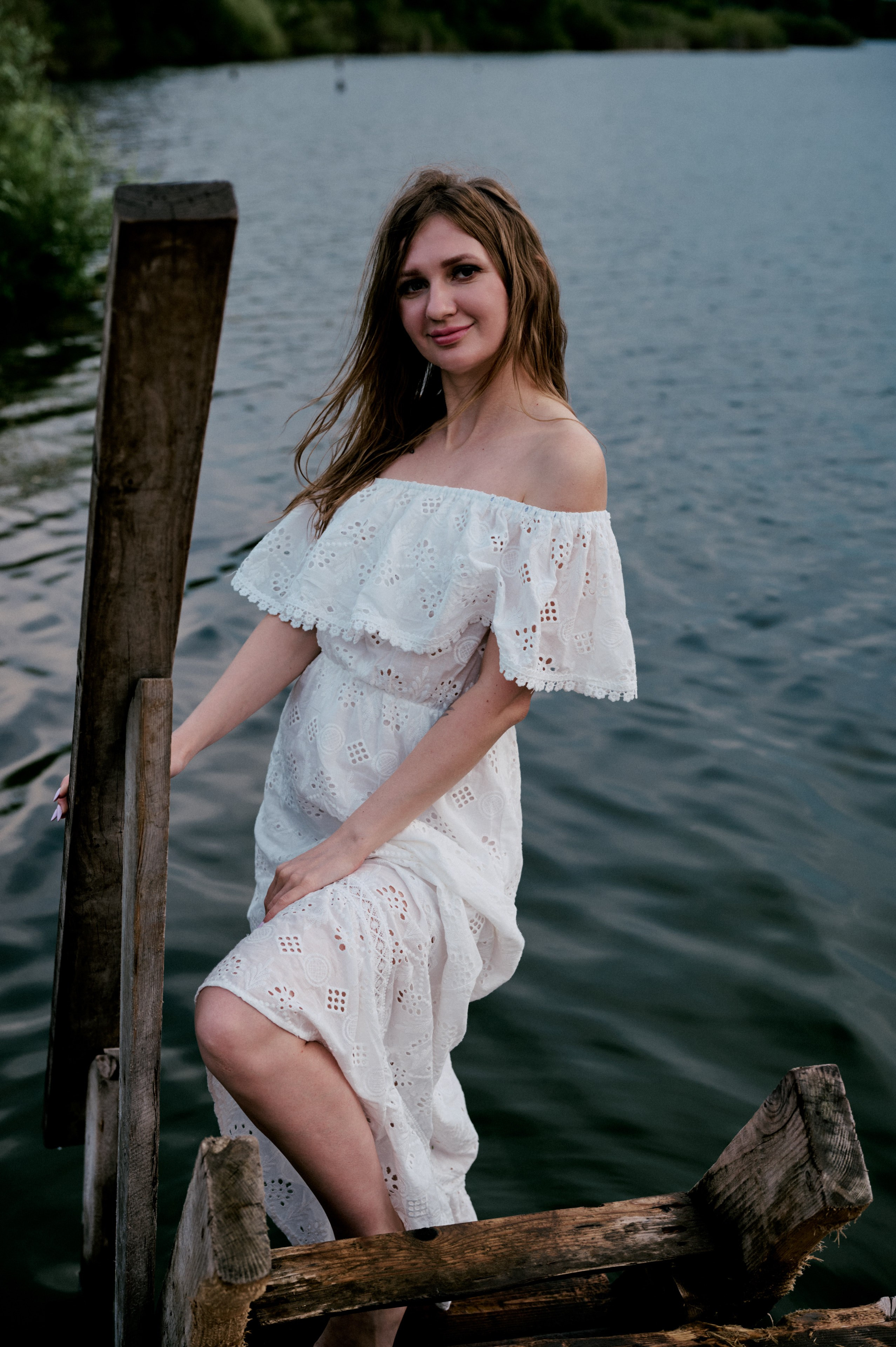Евгения стоит на деревянном пирсе, прислонившись к перилам, в белом платье.
