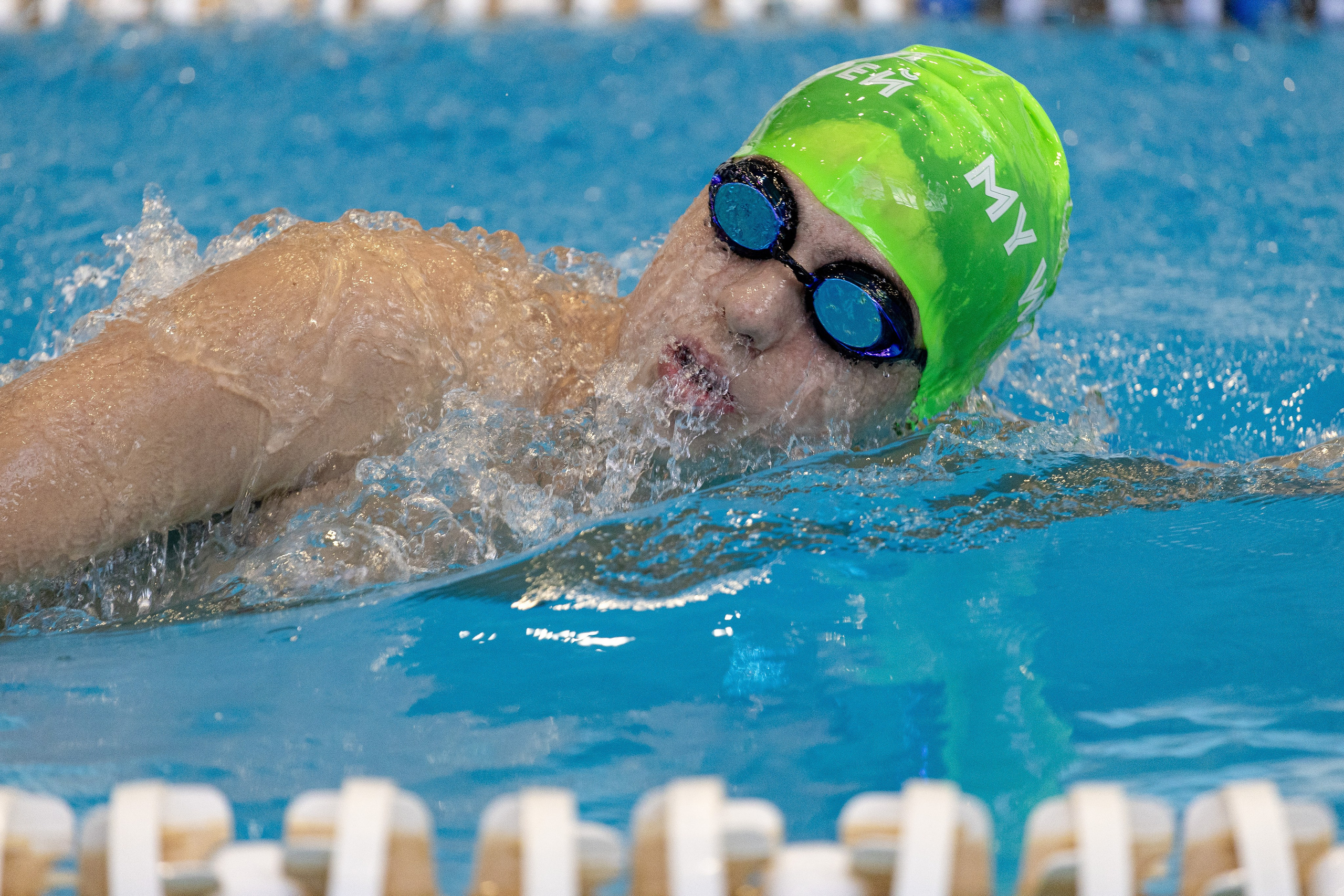 Пловец в зелёной шапочке и очках для плавания выныривает из воды после прыжка с тумбы в бассейн.