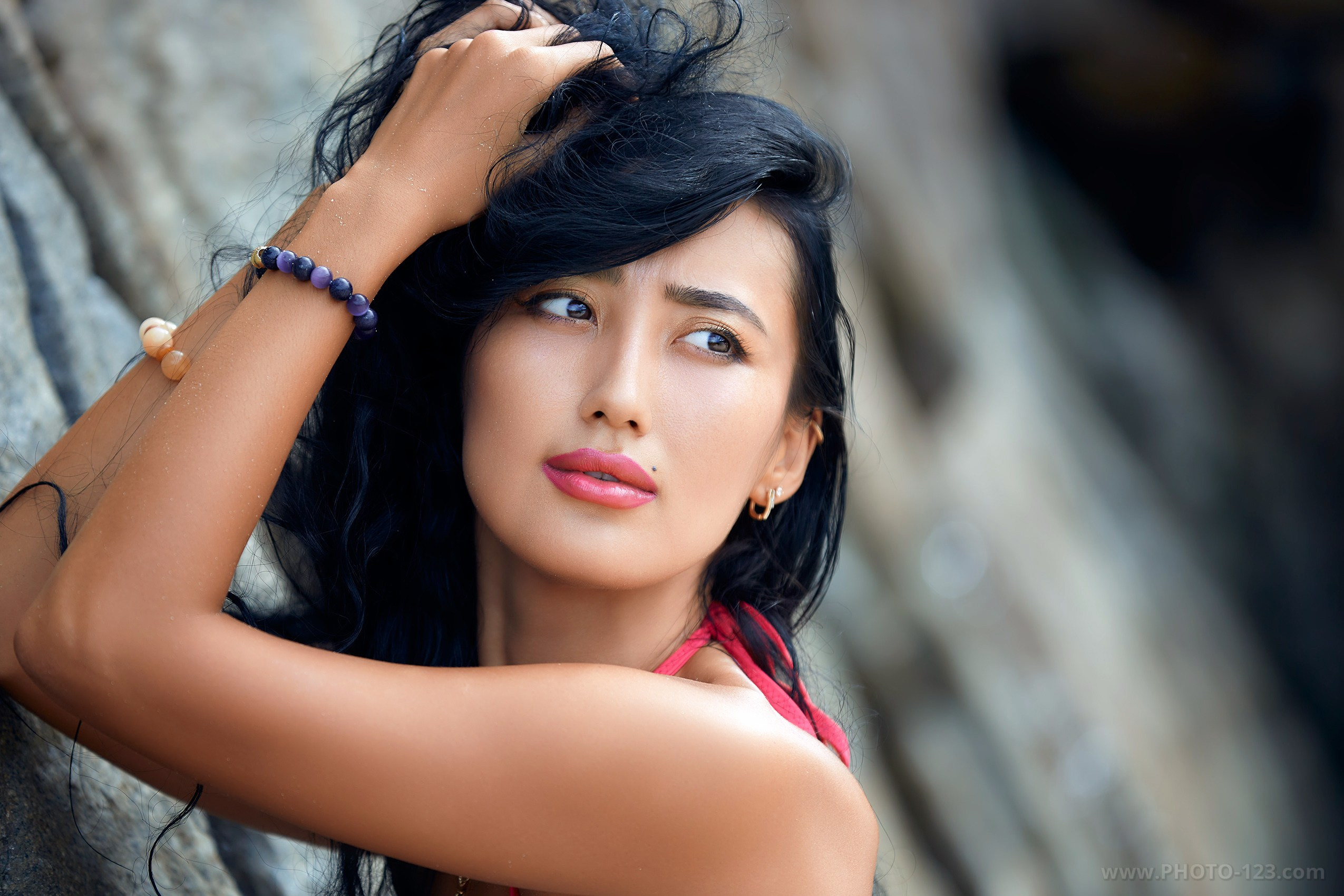Close-up portrait of a young woman with dark hair touching her head, looking to the side, natural makeup with pink lipstick, rocky background, shallow depth of field, emotional outdoor portrait