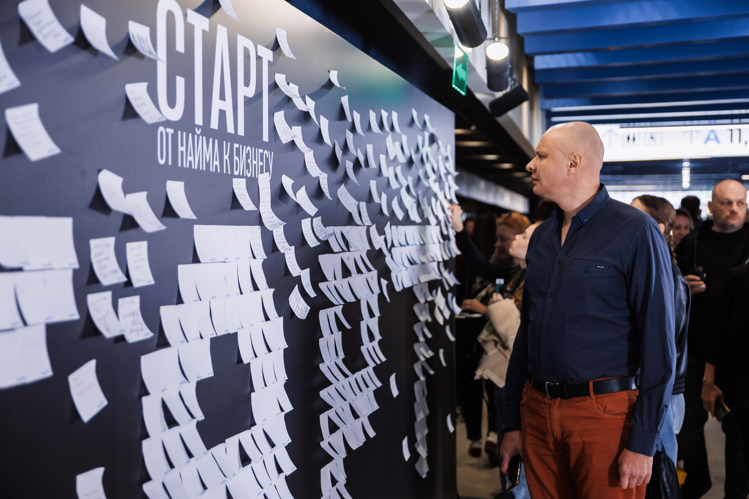 Мужчина в синей рубашке и оранжевых брюках смотрит на большую черную стену, покрытую множеством белых стикеров. Вверху стены написан текст на русском языке. На заднем плане видны несколько человек.