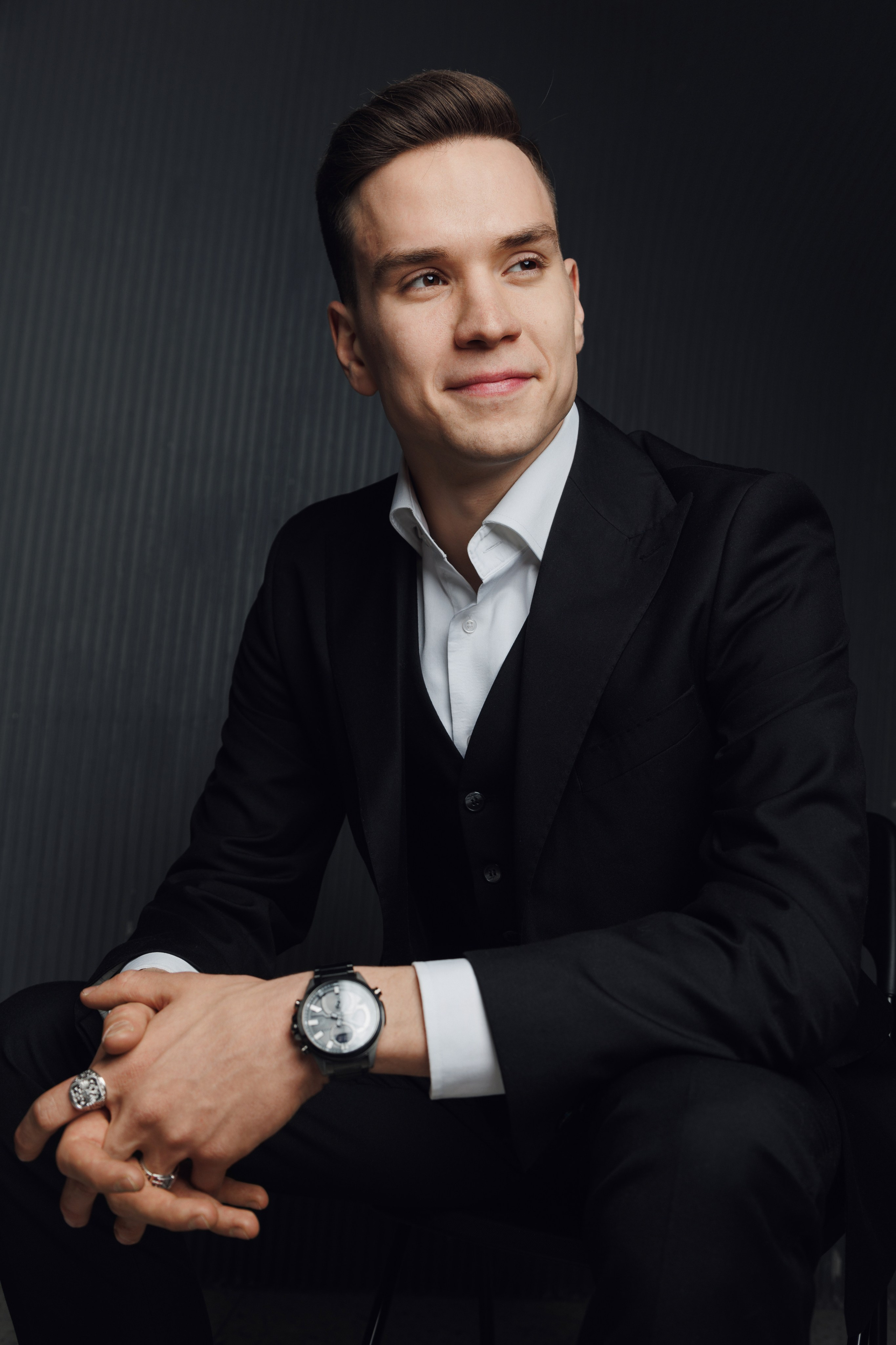 Солидный портрет мужчины в костюме, сидящего на темном студийном фоне.