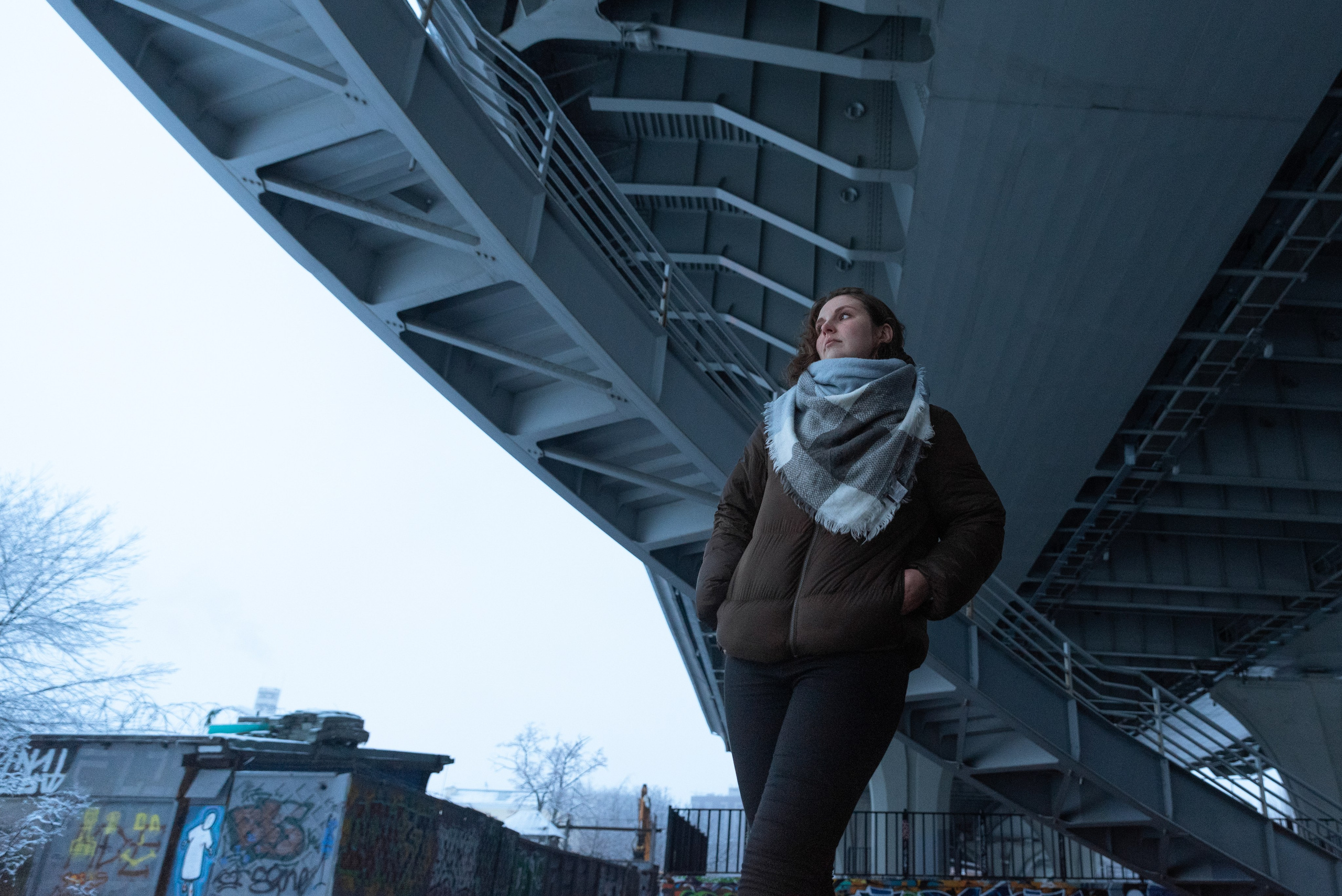 Девушка стоит под мостом. Над ней серые массивные металлические конструкции. На девушке джинсы, широкий шарф и коричневая куртка.