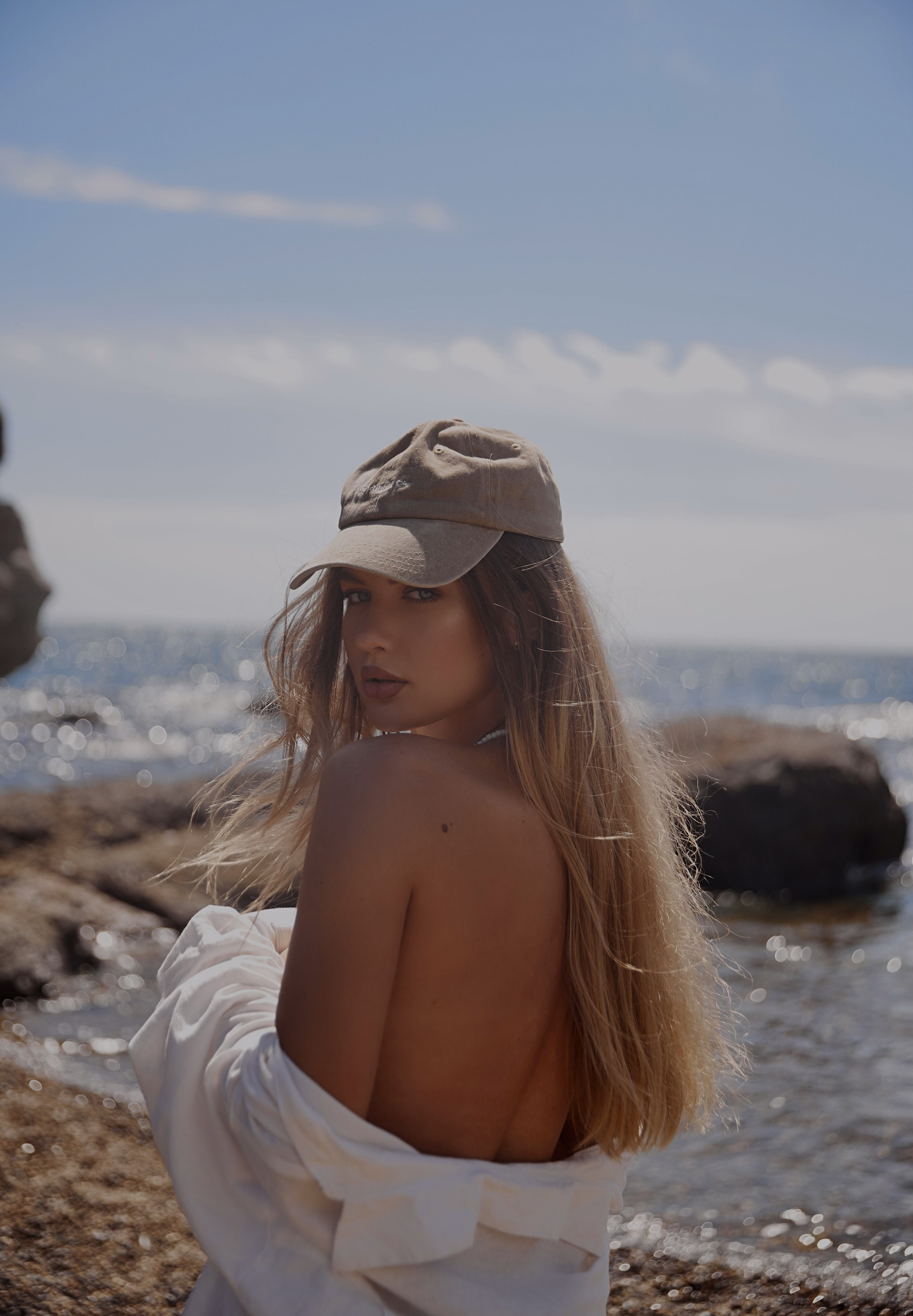 Девушка модель в белой рубашке на голое тело и кепке стоит на берегу