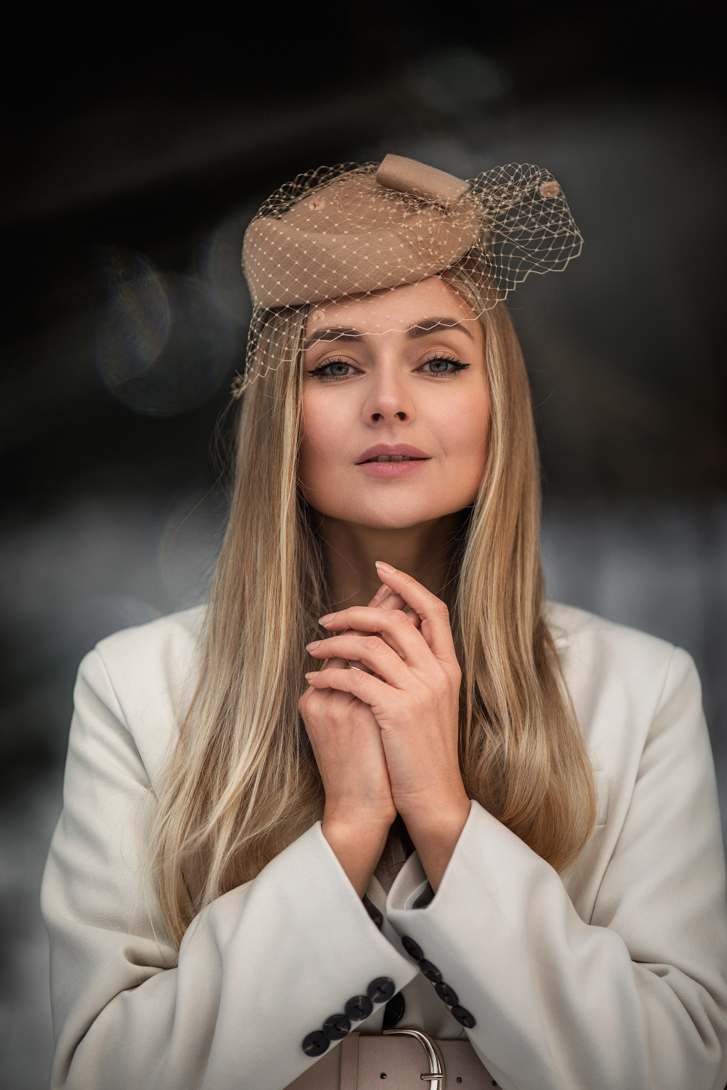 Портрет женщины в бежевой шляпке с вуалью и светлом костюме, мягкий размытый фон.