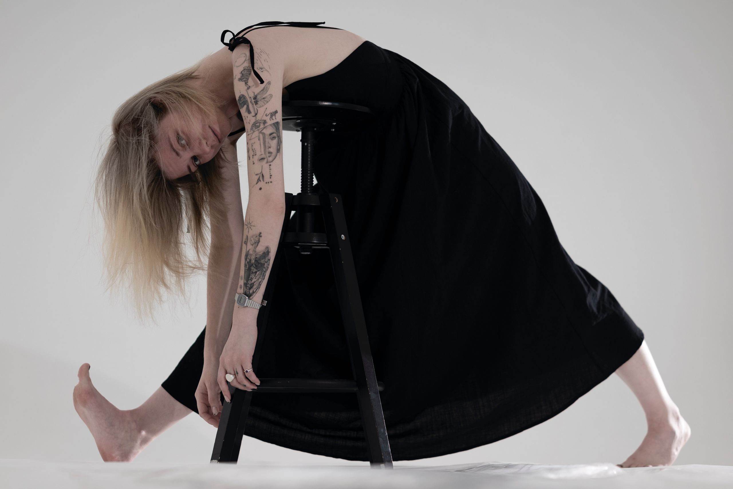 Светловолосая девушка в черном платье наклонилась рядом со стулом