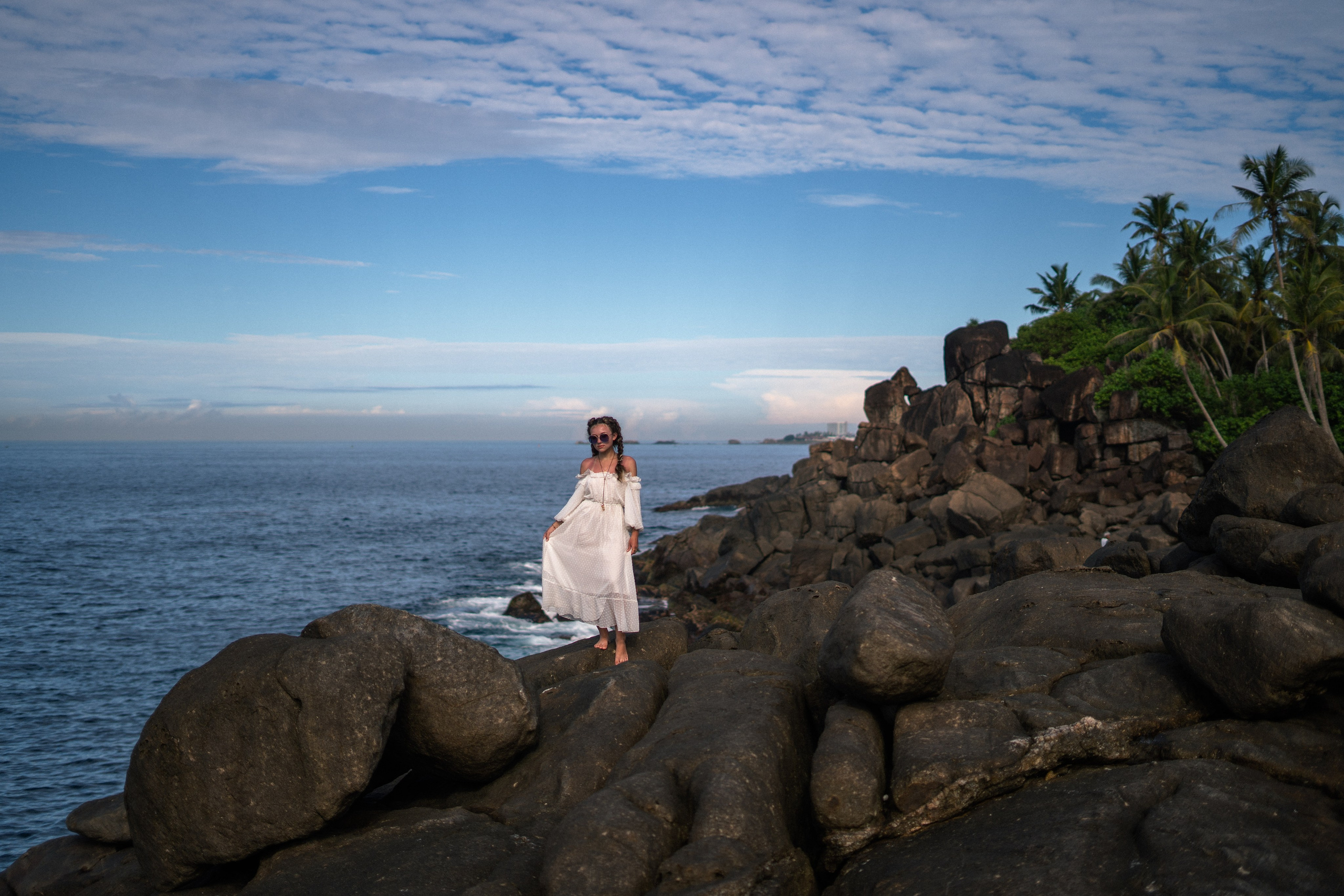 a girl in a white dress and glasses with rocks and the ocean in the background