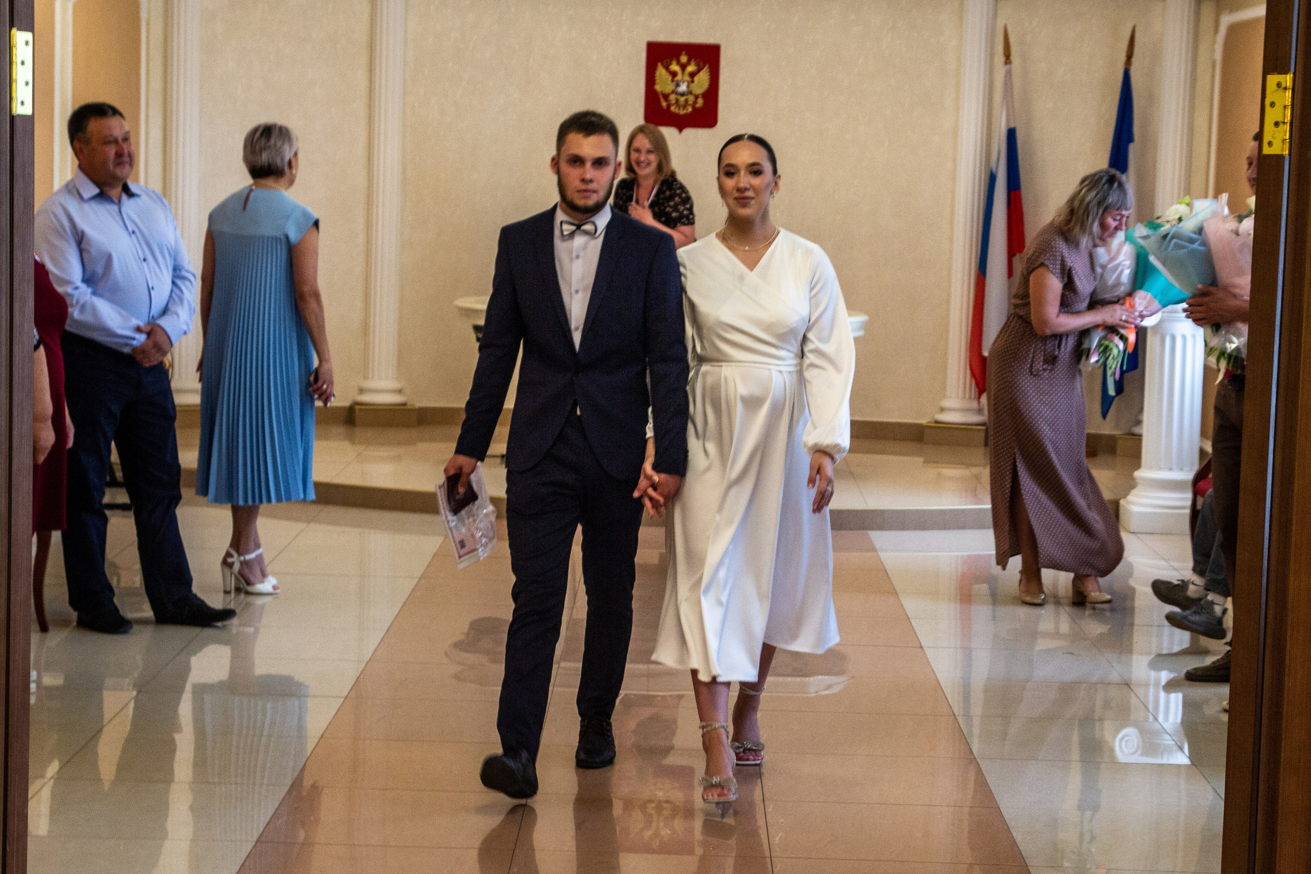 Жених и невеста идут по коридору ЗАГСа после регистрации, гости наблюдают.