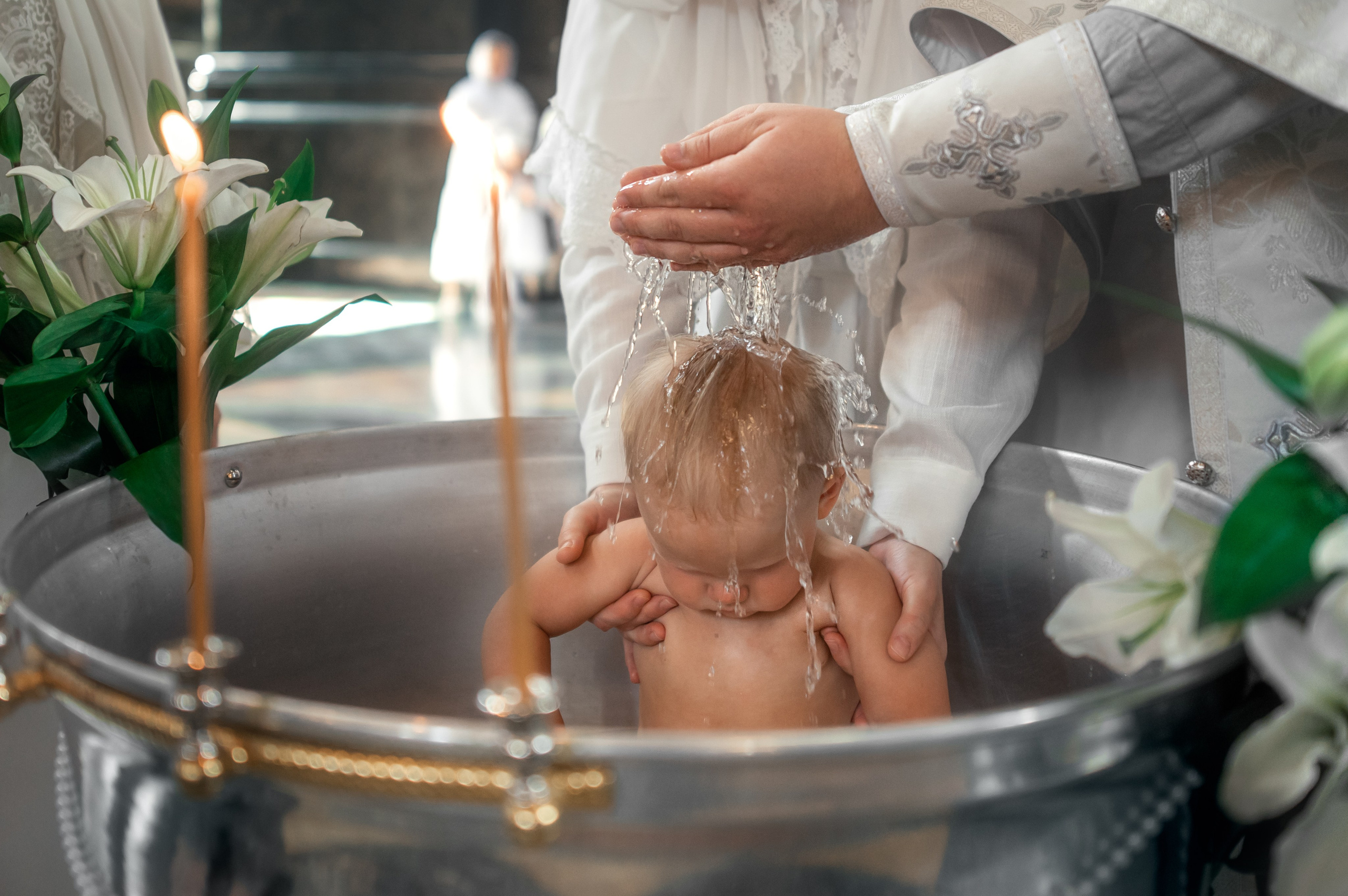 руки священника поливают водой малышку в украшенной лилиями купели