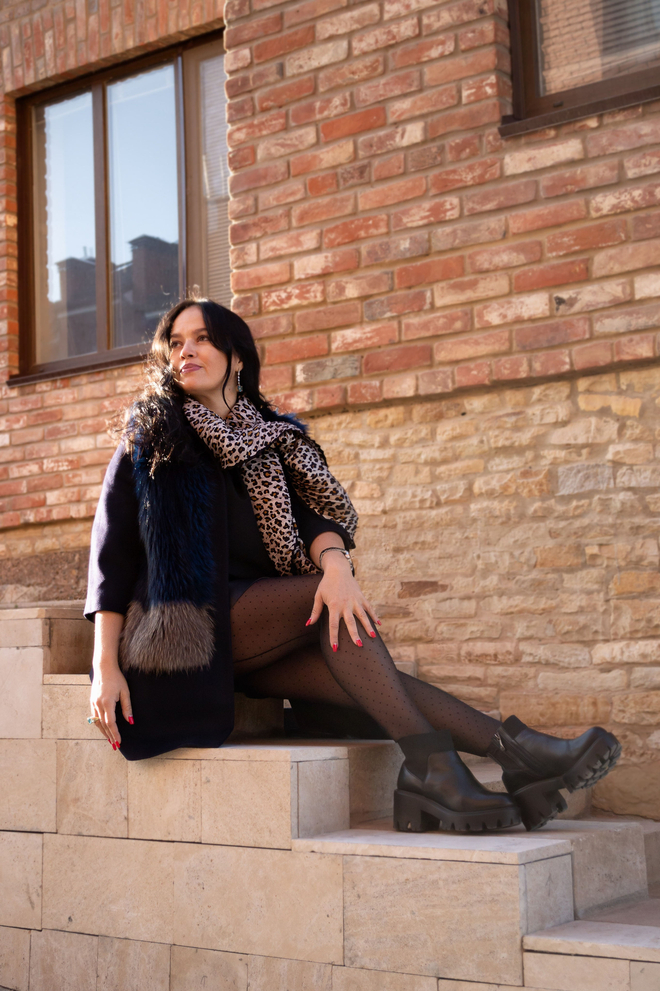 Модно и стильно одетая девушка сидит на ступеньках у кирпичного дома.