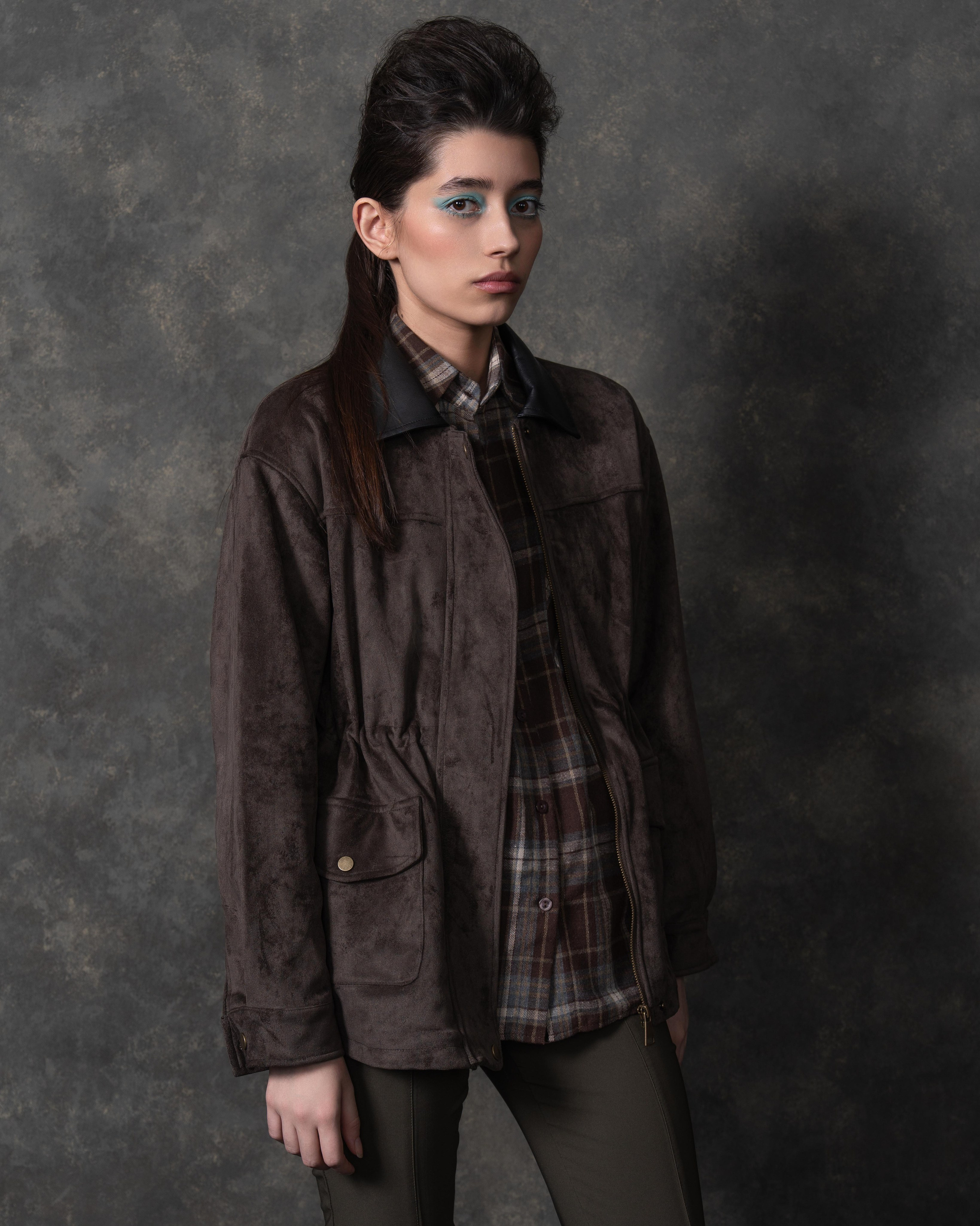 На фотографии изображена женщина, стоящая на фоне серой стены. Она одета в коричневую куртку с меховым воротником и клетчатую рубашку под ней. На ней также зеленые брюки и черные сапоги. В одной руке она держит коричневую шляпу, а в другой — коричневую сумку. Рядом с ней стоит черный чемодан с меховой отделкой и черная ваза. Женщина смотрит прямо в камеру, ее взгляд сосредоточен и уверенный.