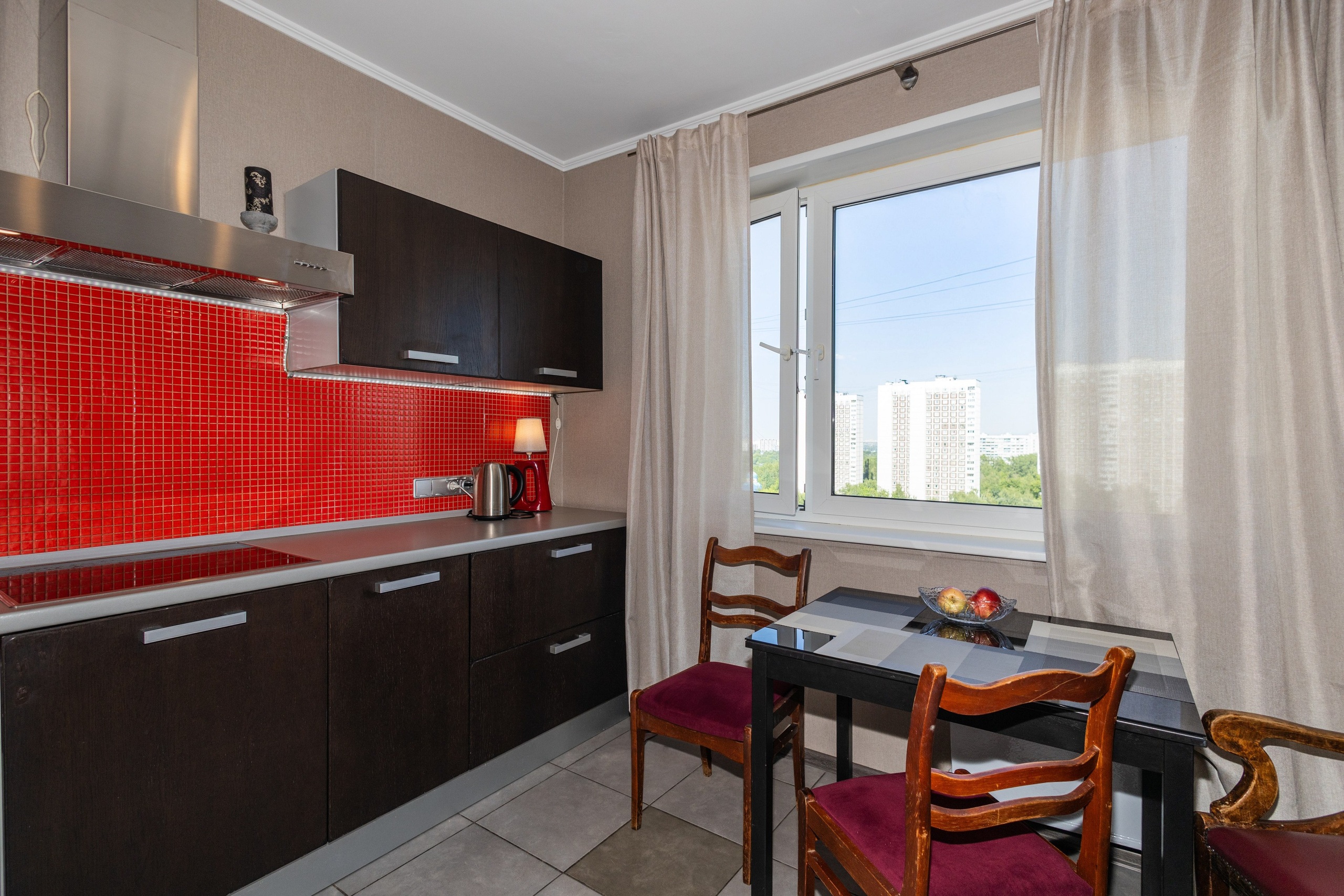 Современная кухня с тёмными фасадами, красным мозаичным фартуком и встроенной техникой; обеденный стол у окна с видом на город, светлые шторы и естественное дневное освещение.