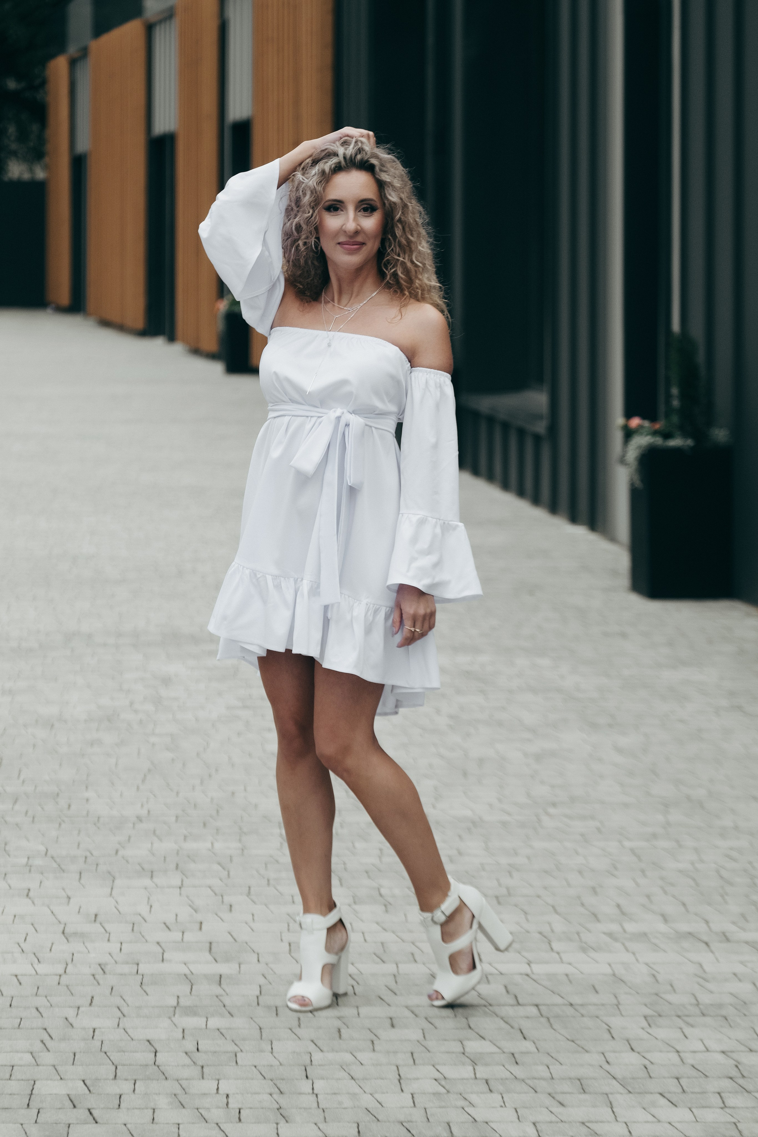 Фотограф в Краснодаре и Архангельске Денис Ожигин. Индивидуальная фотосессия для кудрявой девушки блондинки в городских локациях на фоне кирпичного здания из красного кирпича в белом платье