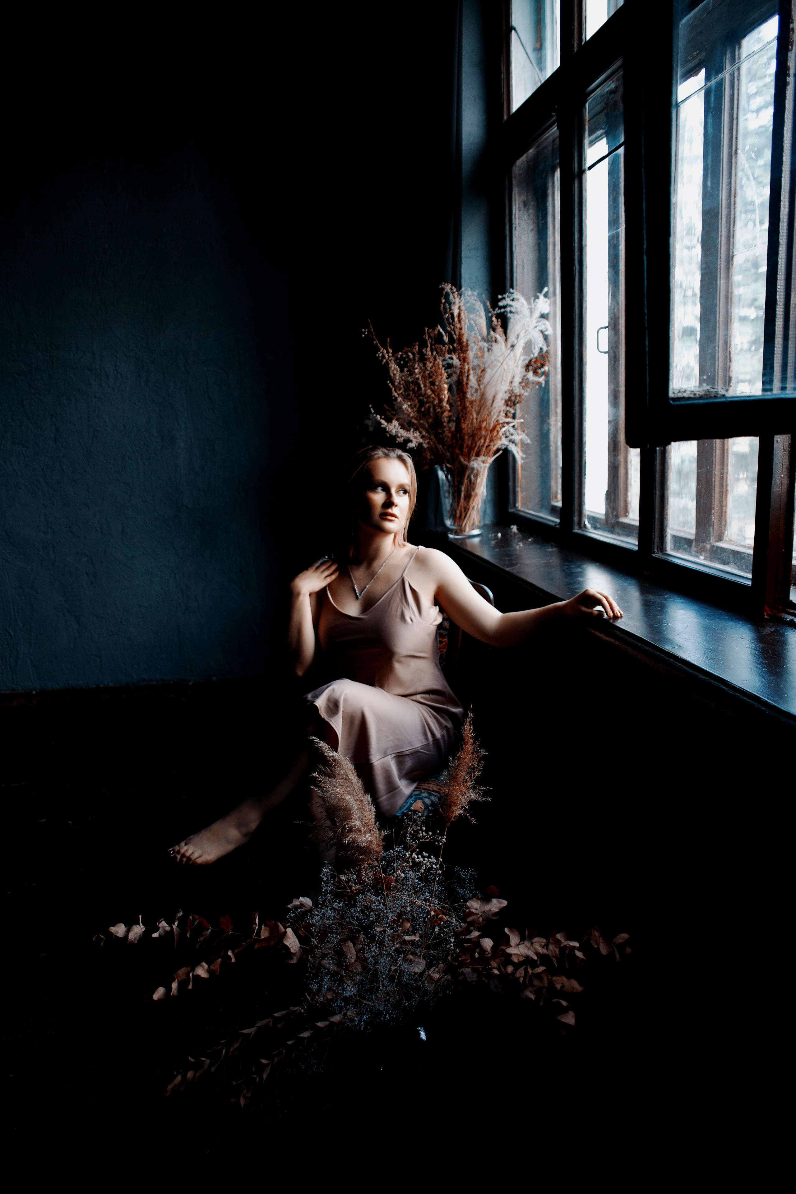 Девушка в платье-комбинации сидит у окна на фоне цветов в тёмной комнате.