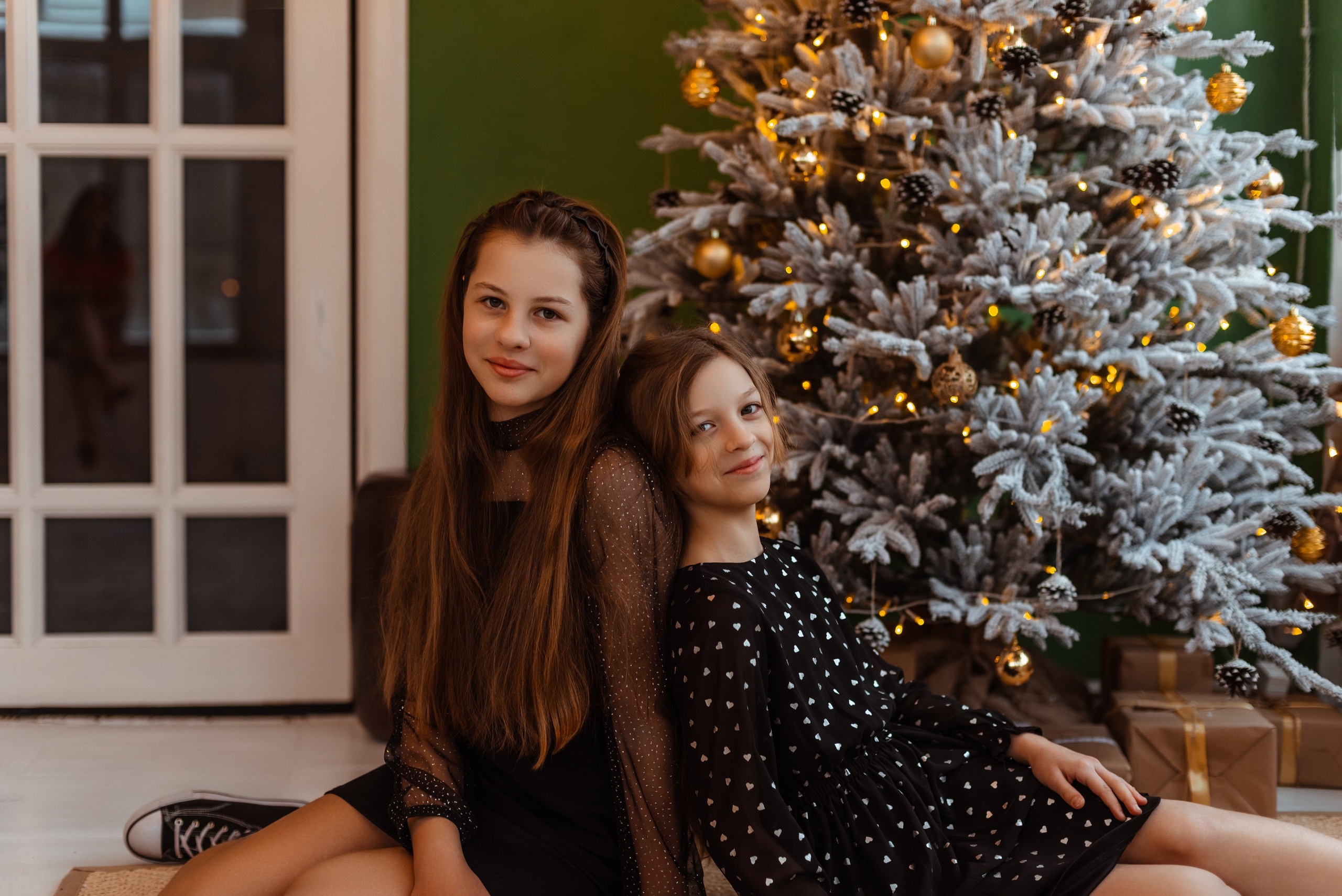 Две сестры вместе улыбаются на камеру, наряженные в праздничные платья, стоя перед яркой новогодней ёлкой