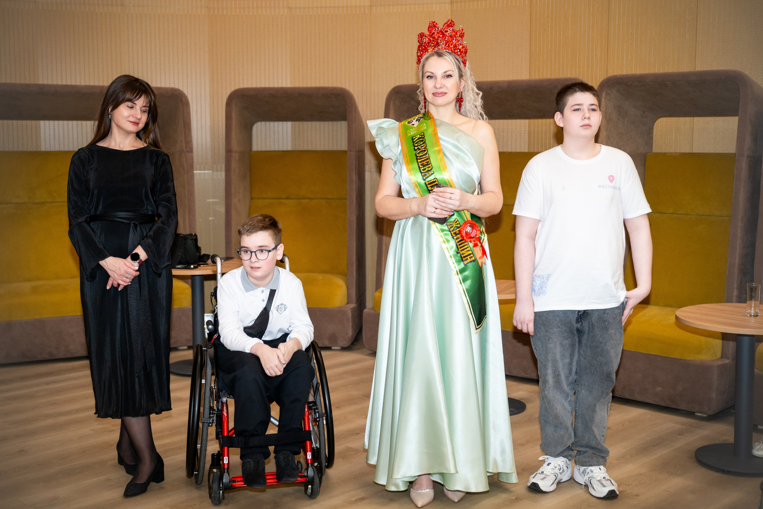 "Королева бала в светло-зелёном платье и короне стоит рядом с мальчиком на инвалидной коляске и другими участниками. Инклюзивный бал «Мы все равны». Фотограф Мальцев Евгений.