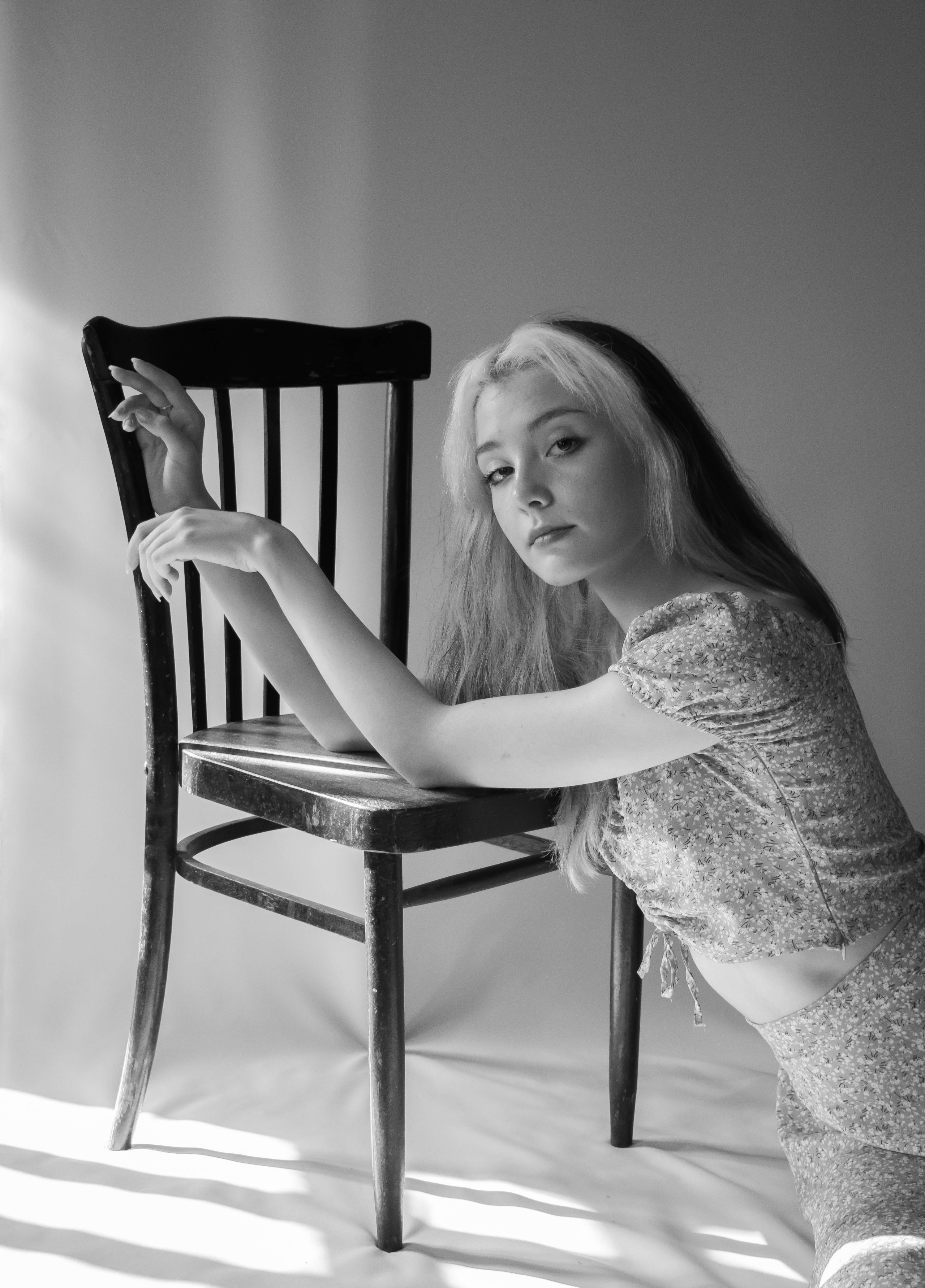 Черно-белая фотография девушки в платье на белом фоне, позирующая со стулом