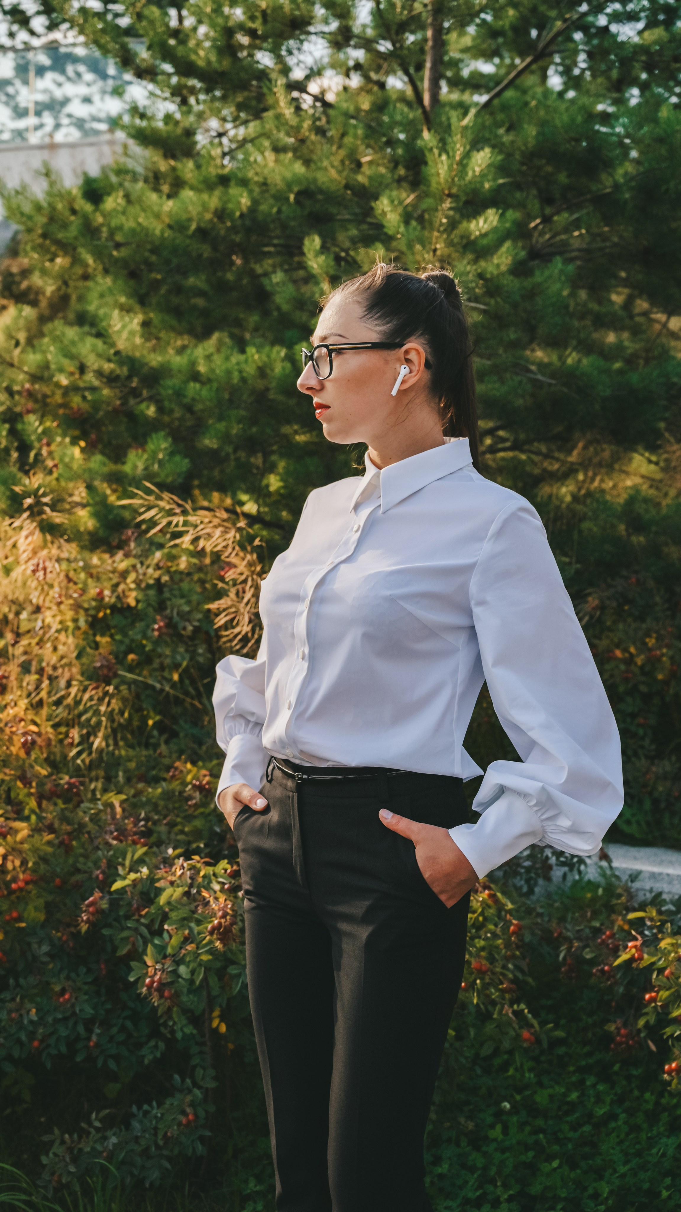 Молодая женщина в деловом и элегантном образе с белой блузкой, черными брюками и черными очками стоит на фоне живописного зеленого ландшафта. Ее длинные волосы аккуратно уложены в высокий хвост, что придает ей современный и профессиональный вид. Наушники в ухе добавляют элемент современности и стиля. Она одета в строгий комплект, который идеально сочетается с окружающей природной средой. Светлый фон неба и яркие краски растений создают ощущение свежести и легкости. Этот снимок демонстрирует мастерство работы с освещением и композицией, делая акцент на игре света и тени, а также на гармоничном сочетании человека и природы. Perfect for business and lifestyle photography enthusiasts!