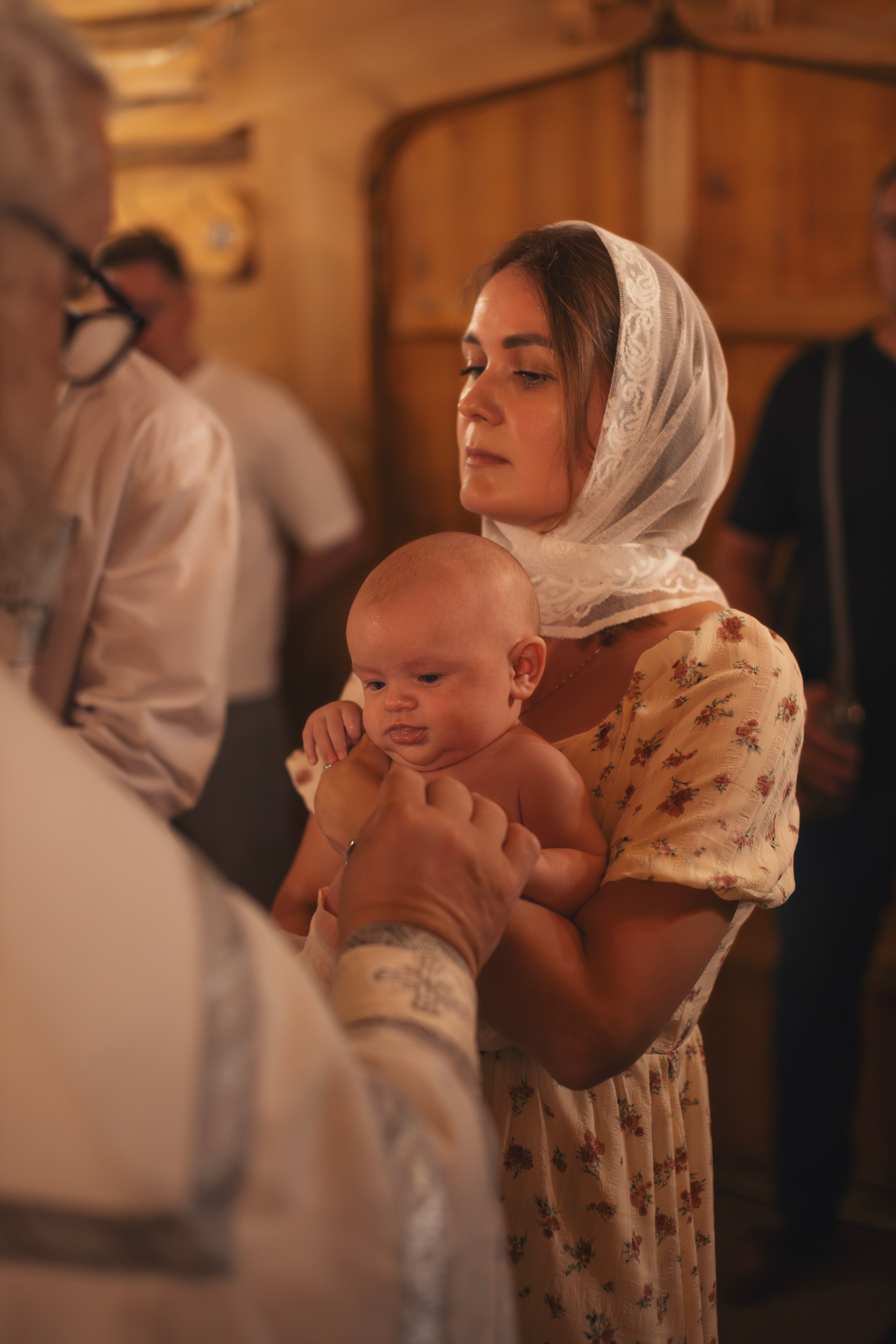 Крестная держит ребенка во время таинства крещения в церкви Успения Пресвятой Богородицы, священник проводит обряд. Крестная и ребенок сосредоточены на моменте священнодействия.