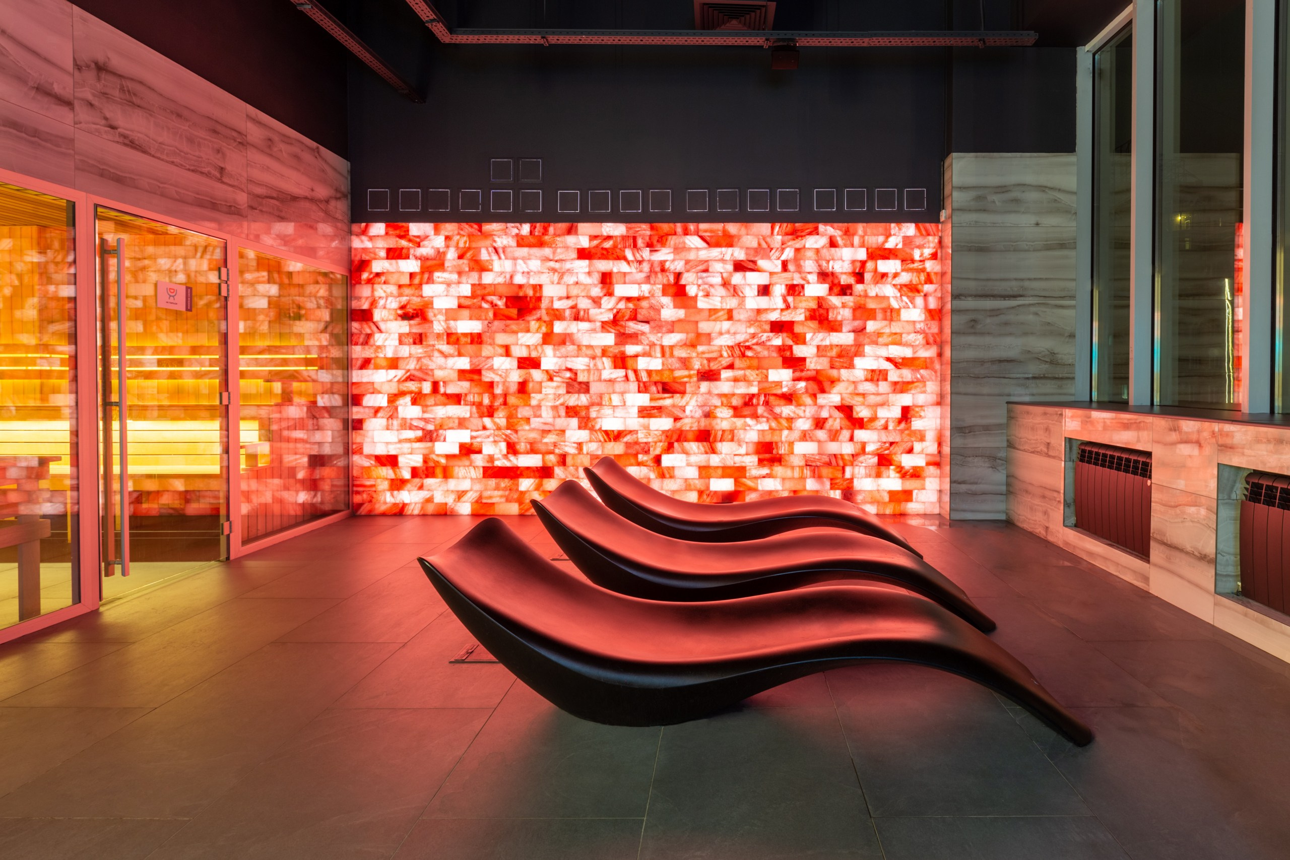 Фото в красных тонах. Сауна с солевой стеной и красивыми лежаками. Подсветка в сауне. Баня сауна в тренажёрном зале фитнес клуба.