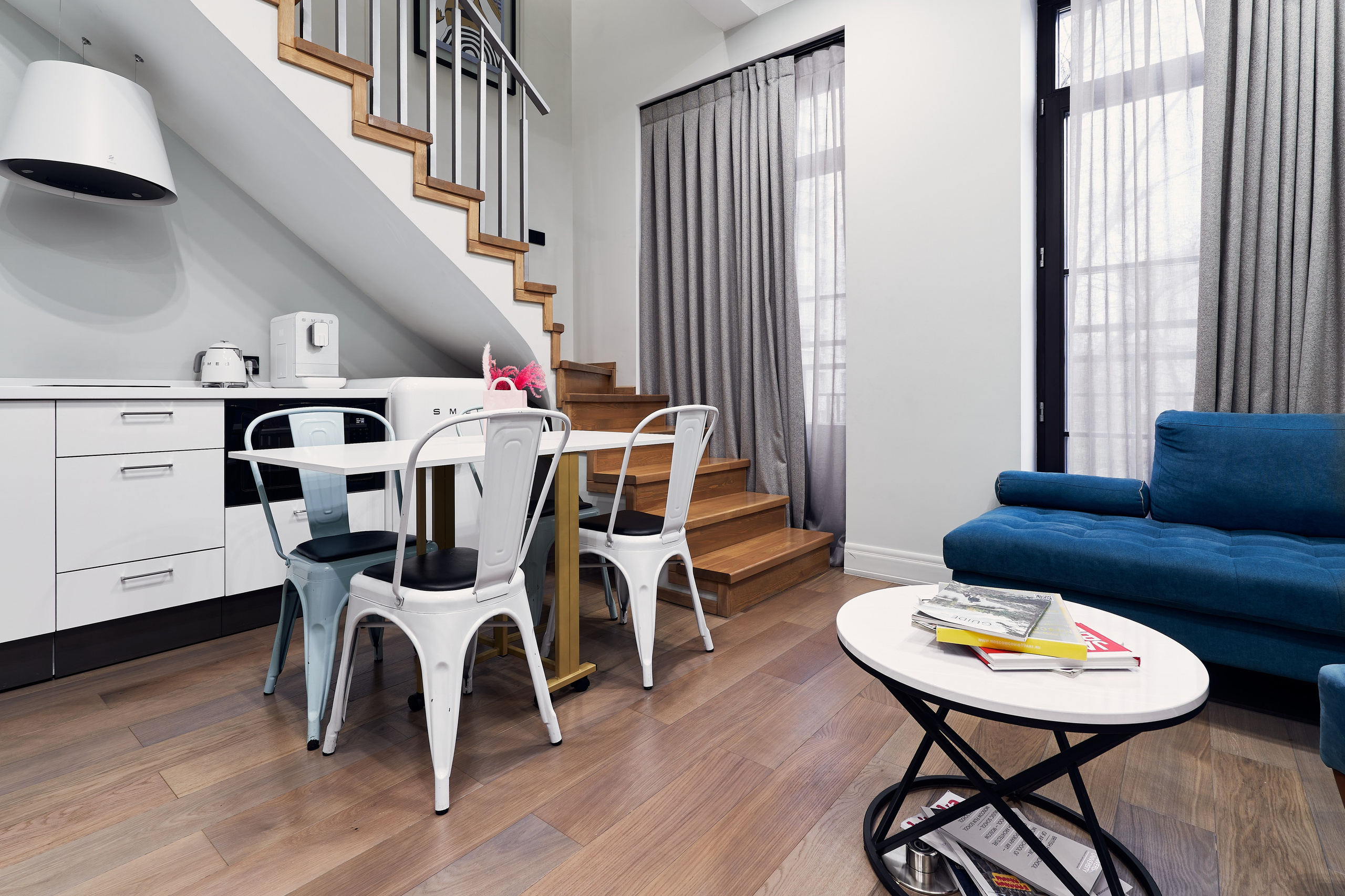 Интерьерная фотосъемка гостиной в московских апартаментах с диваном, кухонной зоной, журнальным и обеденным столами, двумя окнами и витой лестницей на 2-й этаж 