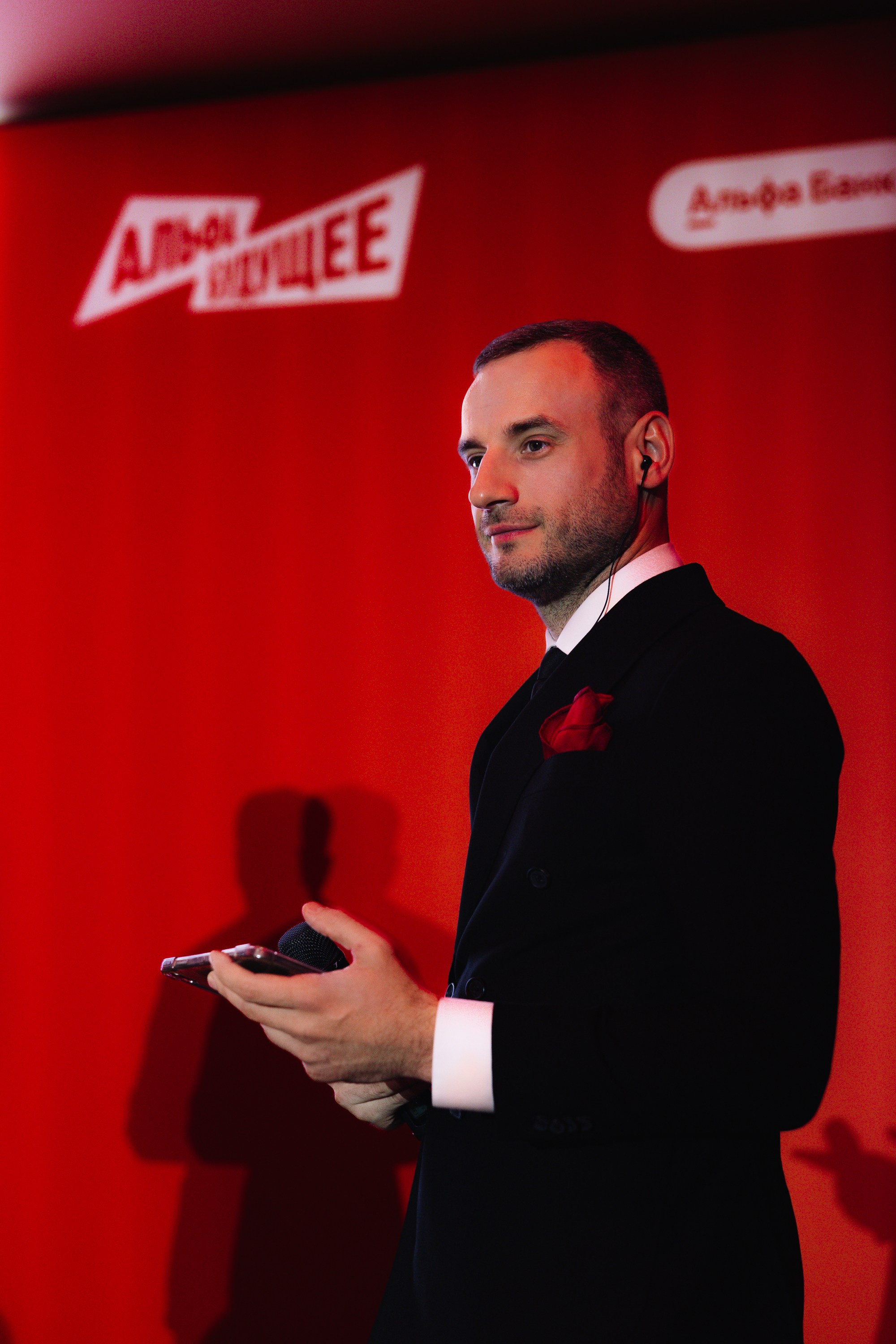 Портрет спикера: Крупный план ведущего или организатора в черном пиджаке на фоне пресс-волла.