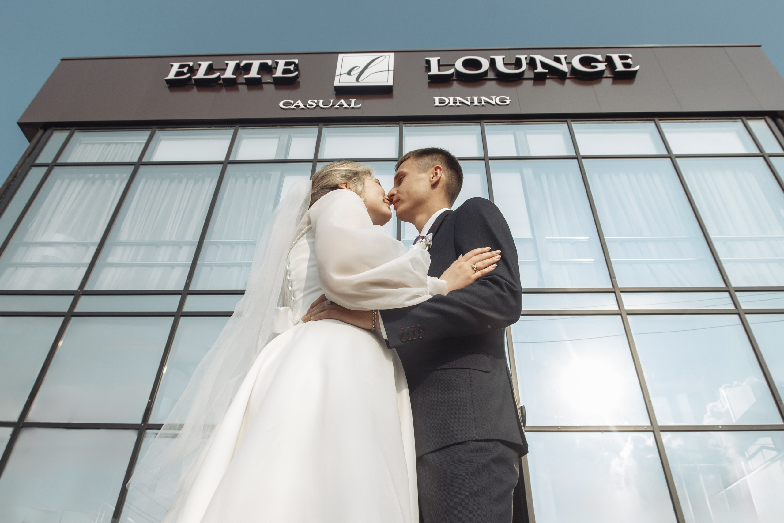 Пара целуется на фоне современного здания с надписью «Elite Lounge» — свадебный поцелуй на фоне архитектуры.