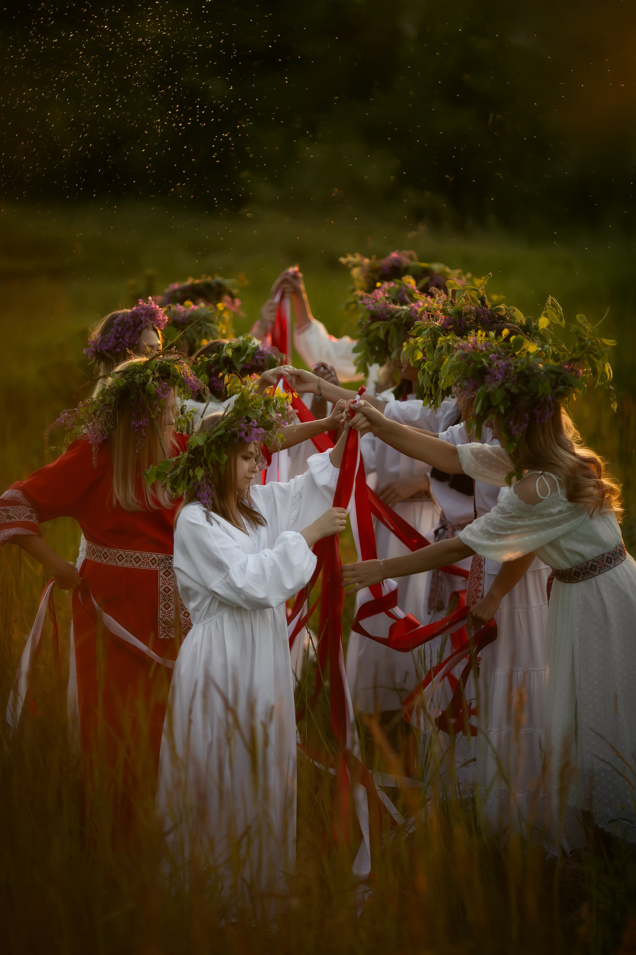 Группа девушек в венках из трав и цветов переплетают красные ленты на закате, в центре кадра мягкий тёплый свет, создающий ощущение праздника и единства.
