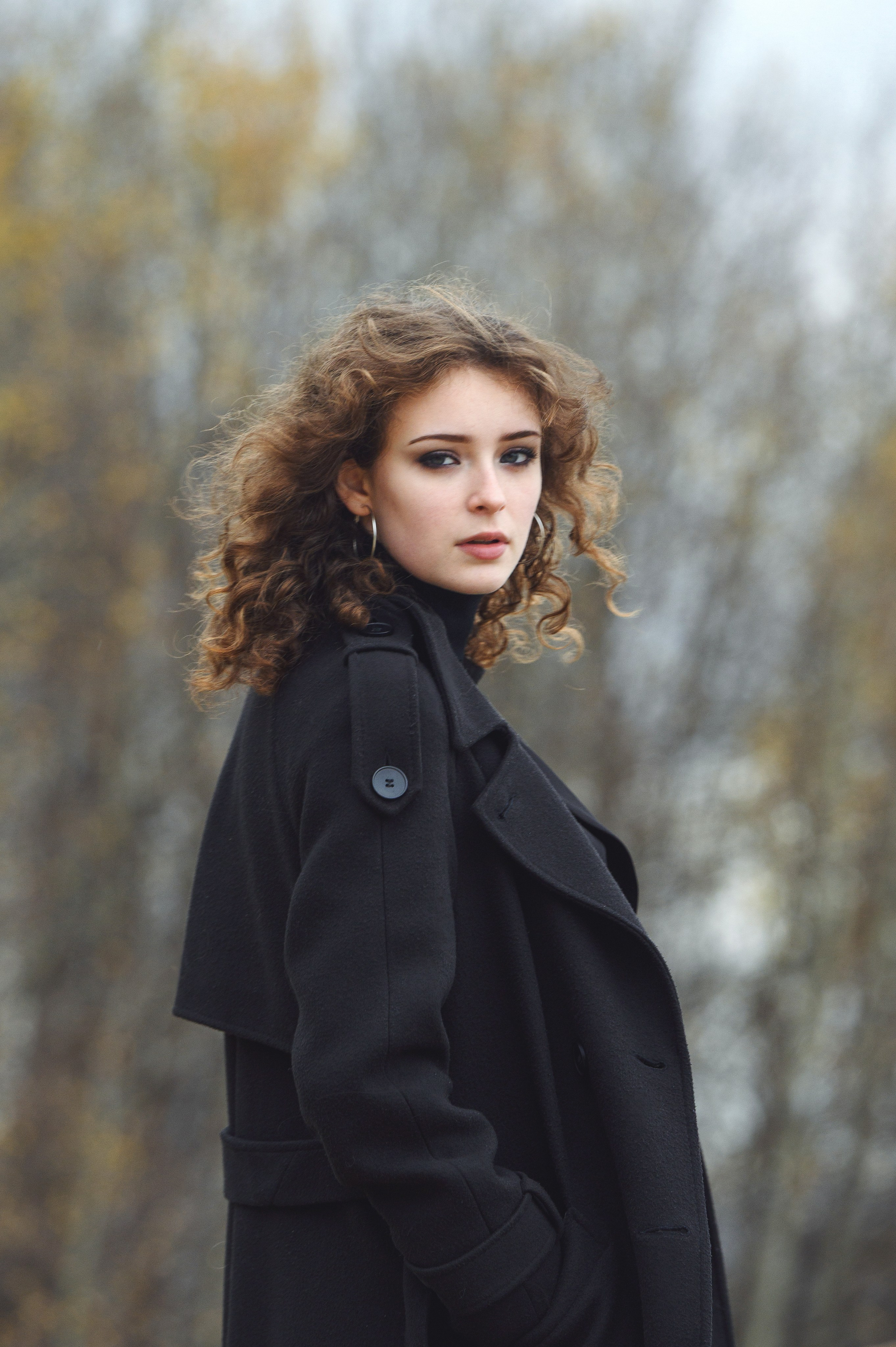 Портрет кудрявой старшеклассницы в чёрном пальто на мягко размытом осеннем фоне.
