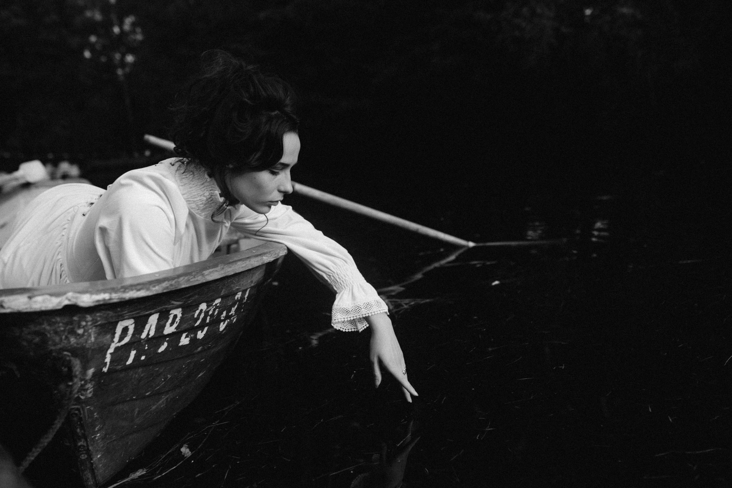 Черноволосая девушка в белом платье с длинными рукавами трогает воду, сидя в лодке