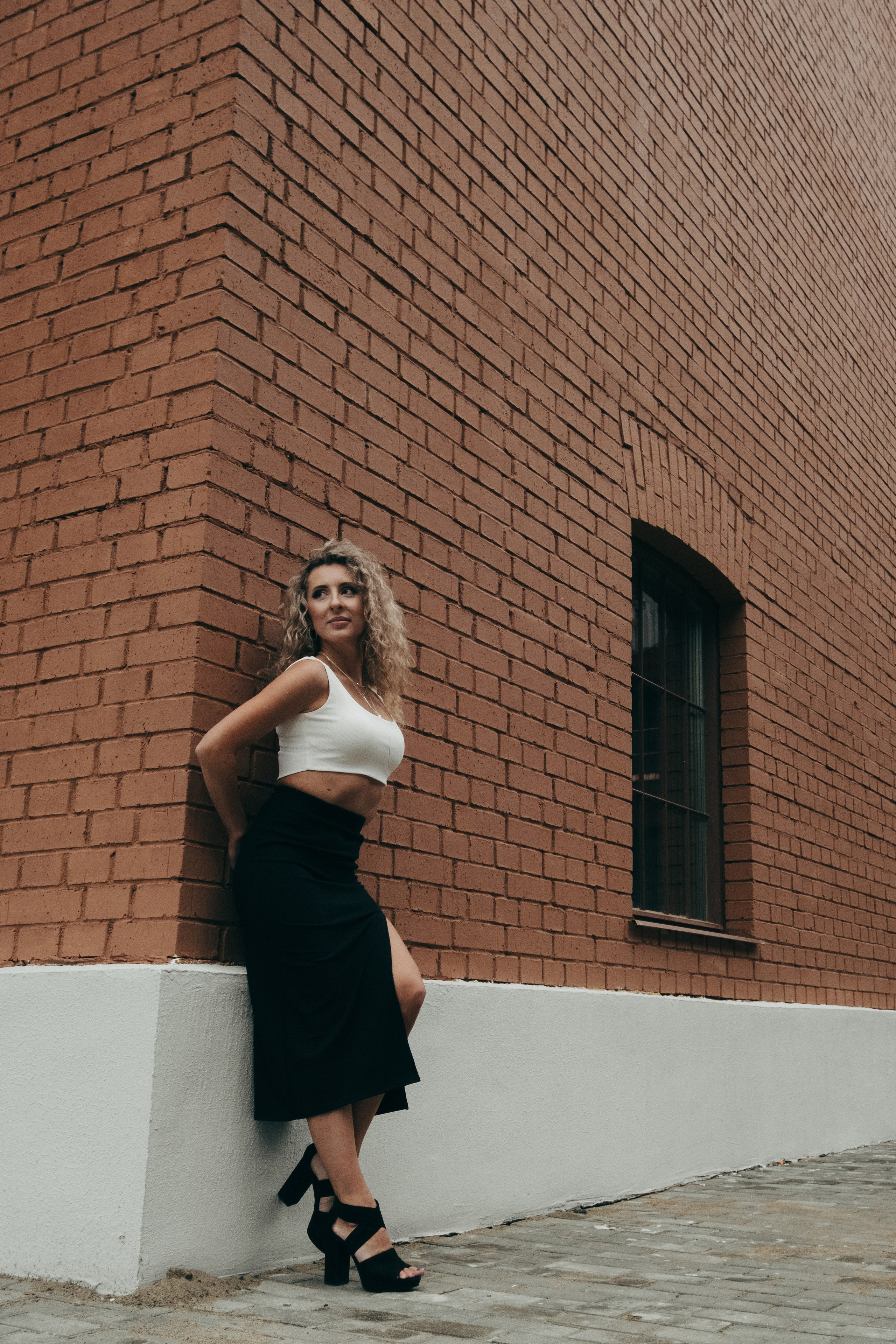 Фотограф в Краснодаре и Архангельске Денис Ожигин. Индивидуальная фотосессия для кудрявой девушки блондинки в городских локациях на фоне кирпичного здания из красного кирпича в белом платье