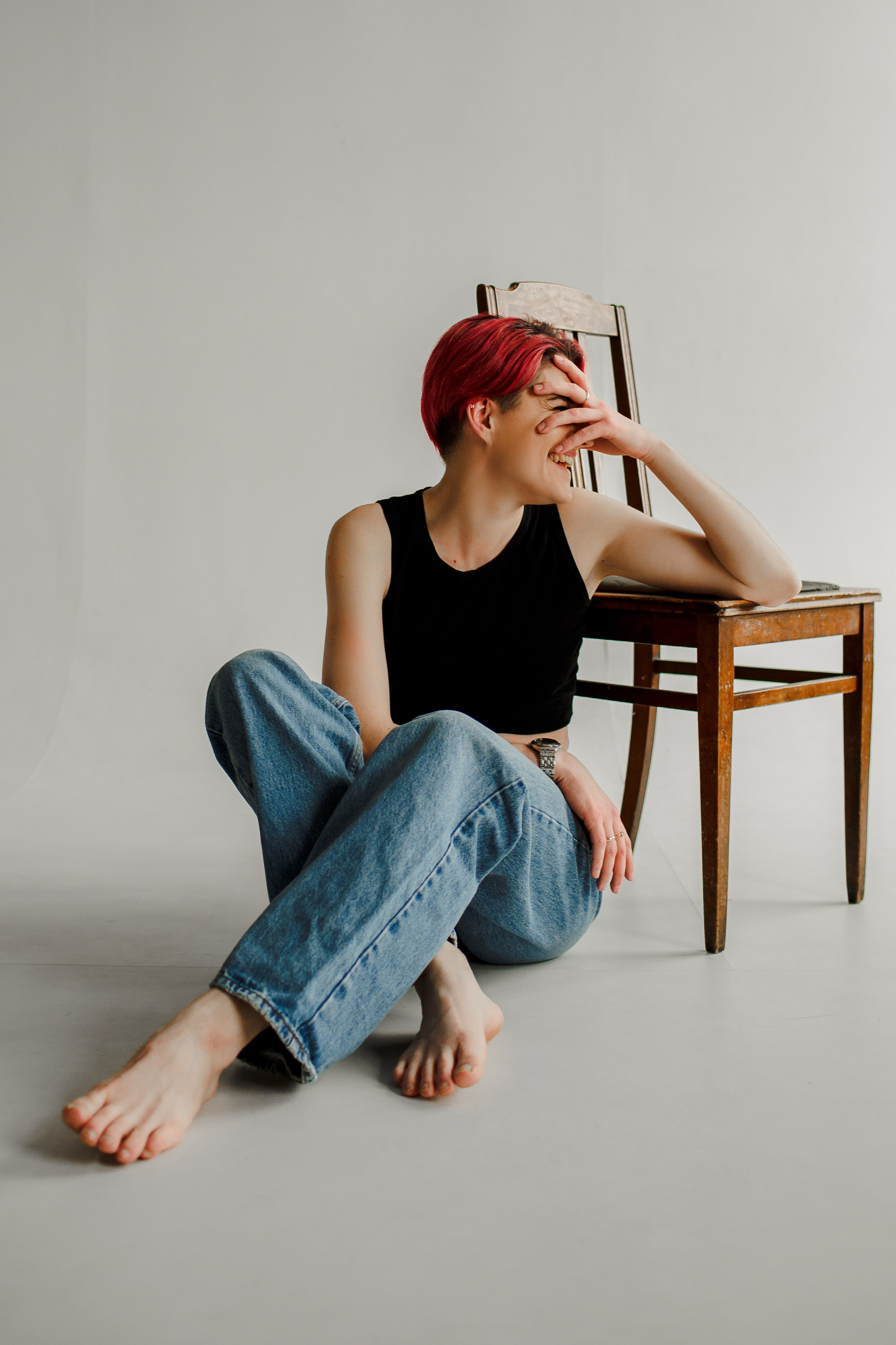 Женщина в джинсах и чёрном топе позирует на деревянном стуле в студии с белыми стенами, её поза динамична, а лицо выражает радость.
