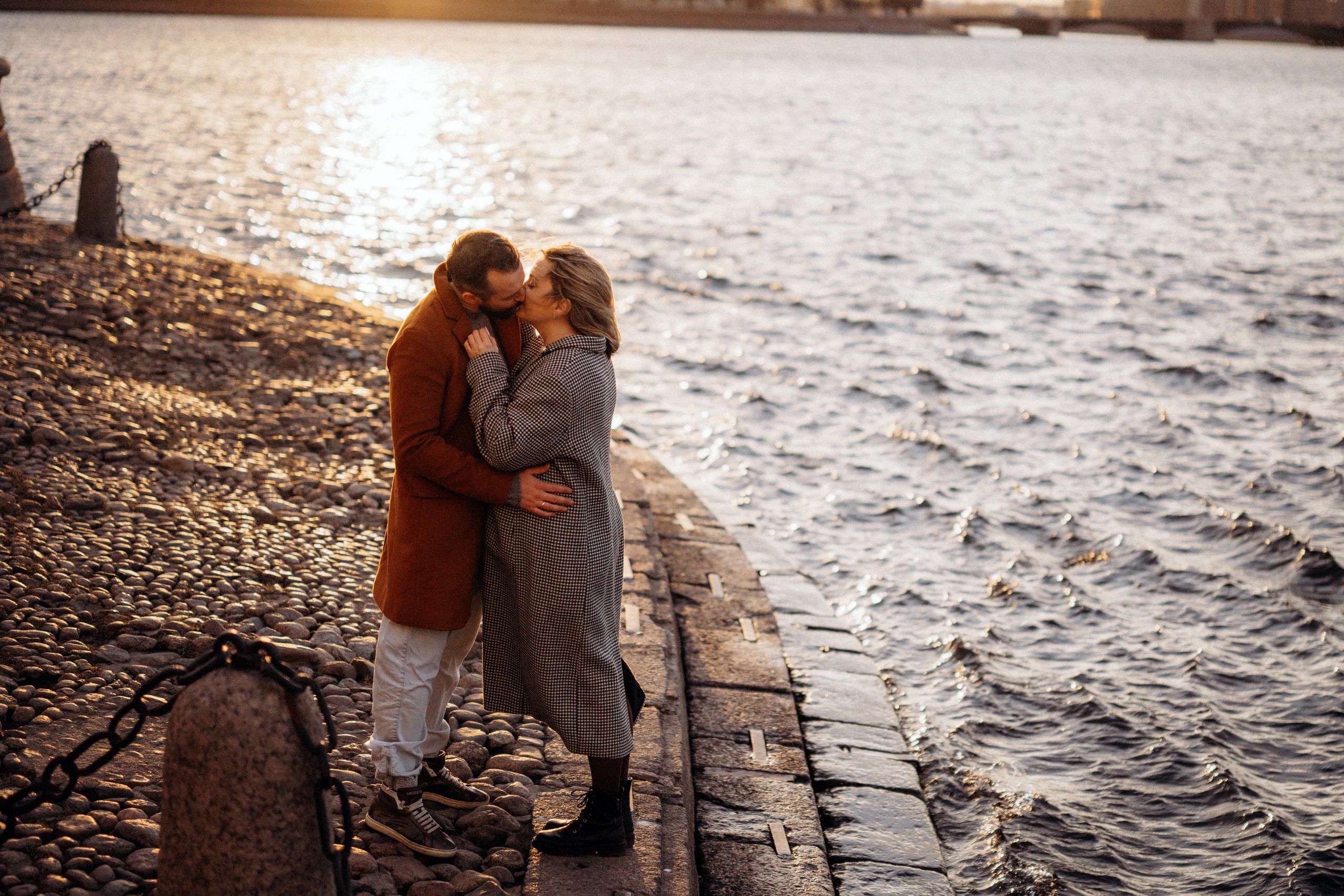 Лавстори Love Story в Санкт-Петербурге у набережной Невы на закате. Пара в коричневых пальто целуется на гранитной набережной