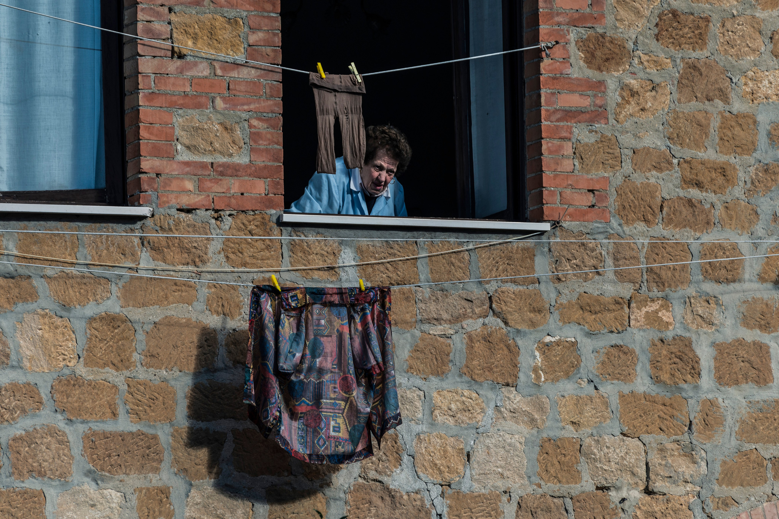 Женщина выглядывает из окна старого каменного здания в Италии; на веревке сушится белье, включая брюки и юбку с узором.