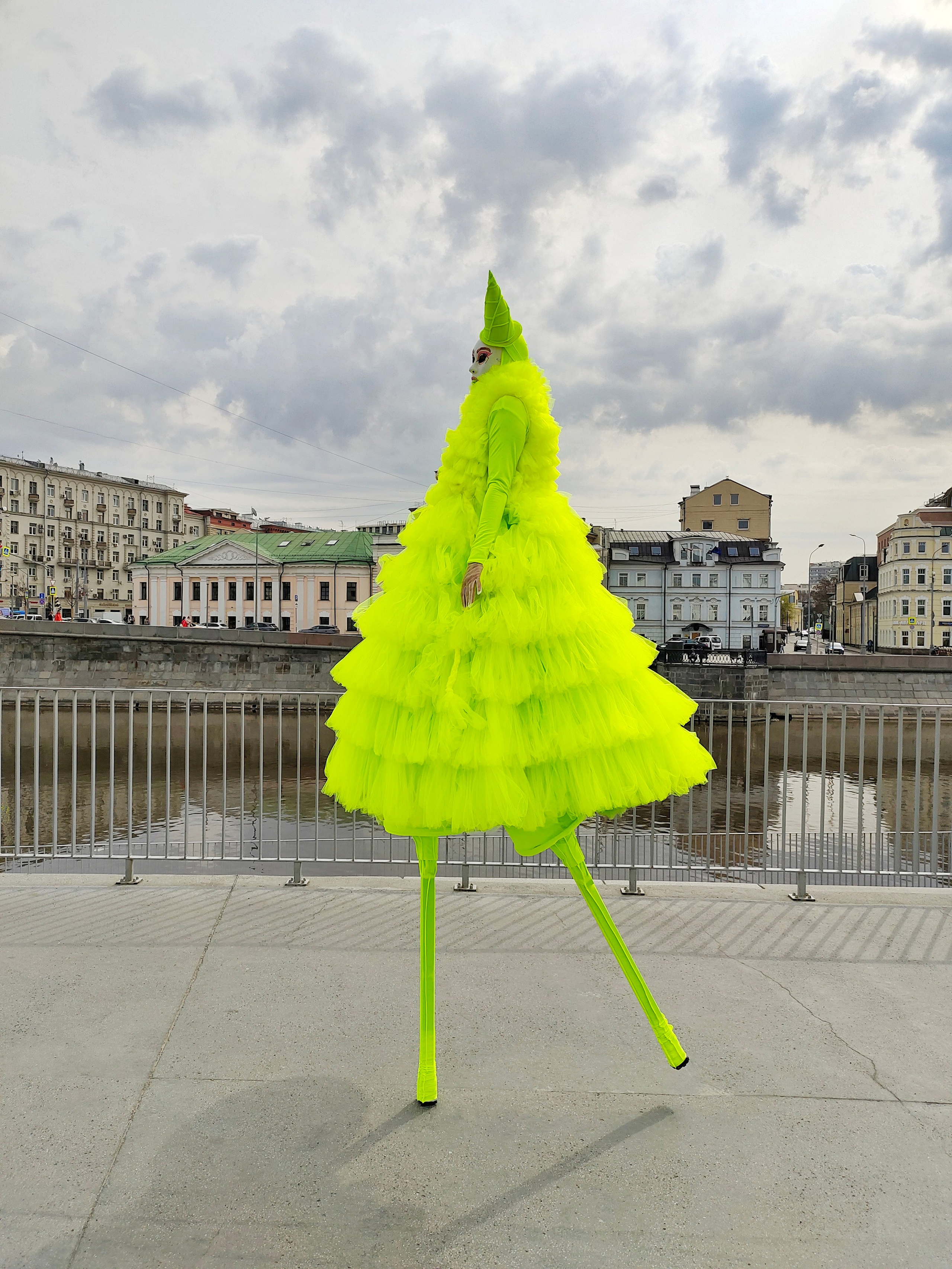 Мим на ходулях в платье лимонного цвета идёт вдоль набережной реки Москва