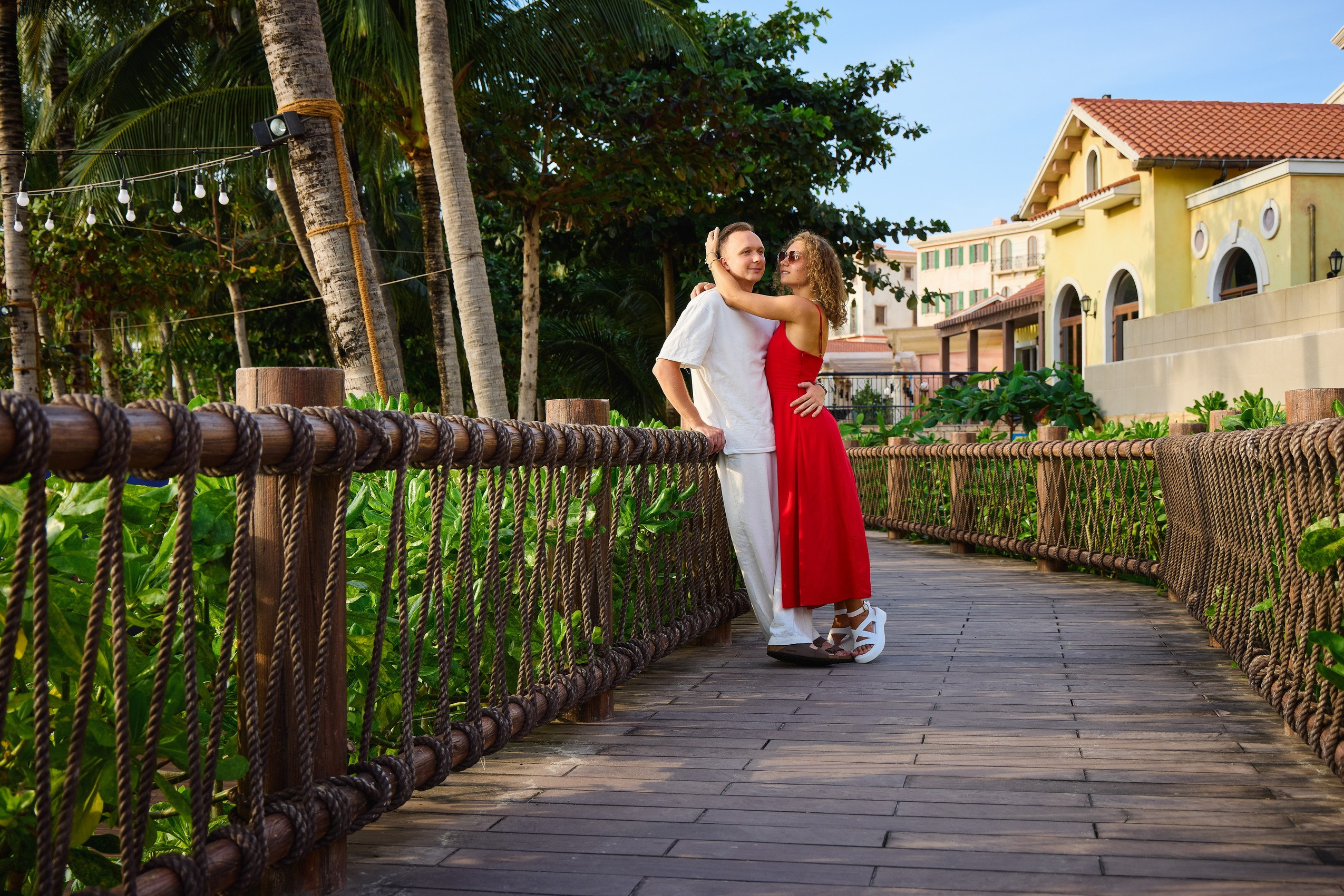 Couple in red and white clothes standing on a wooden boardwalk with rope railings surrounded by palm trees at a luxury resort in Phu Quoc, Vietnam.