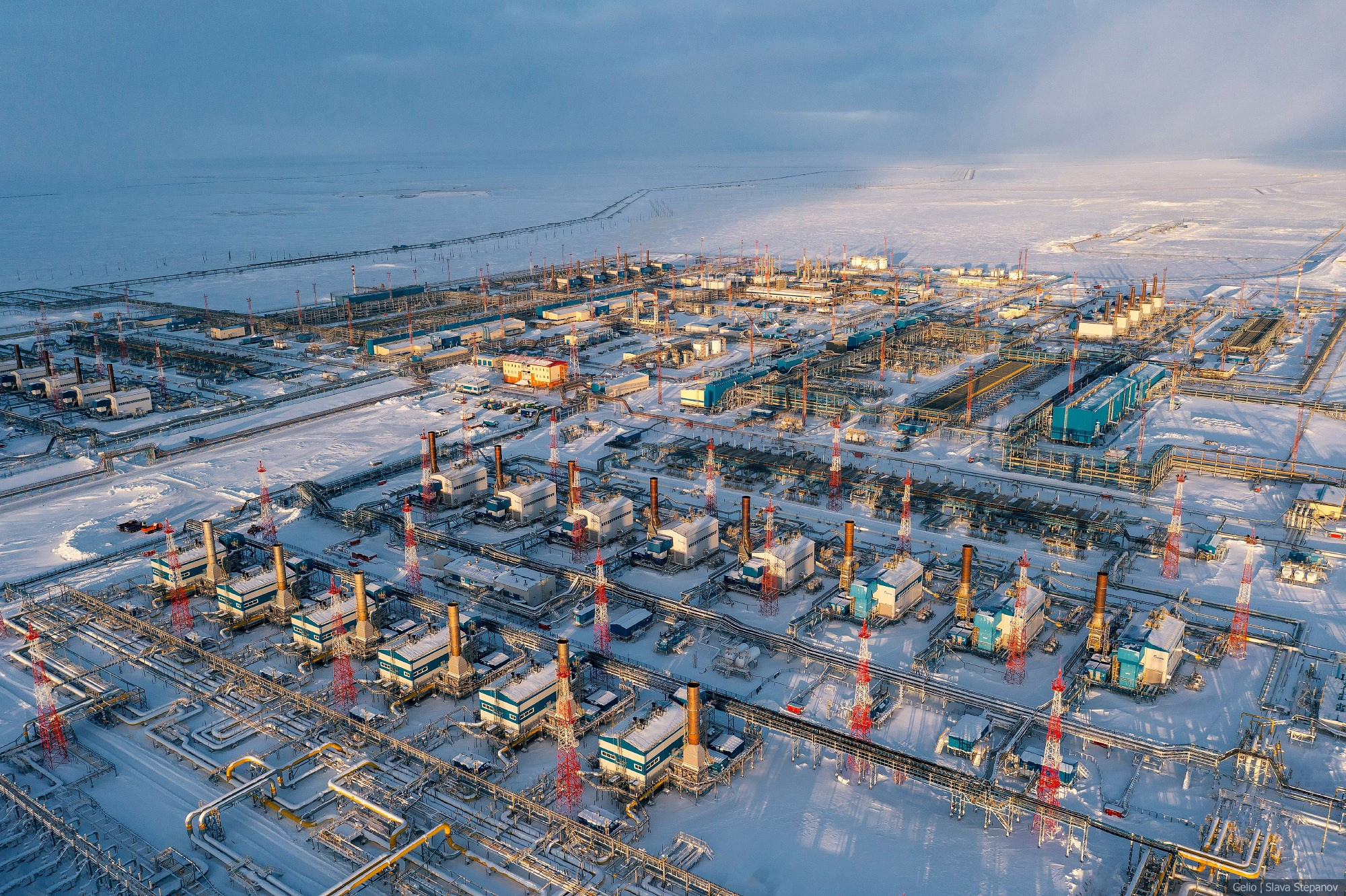 Заснеженная газокомпрессорная станция с высоты, ряд крупных производственных цехов с высокими трубами и сложная сеть зимних трубопроводов на северном месторождении, Gelio.