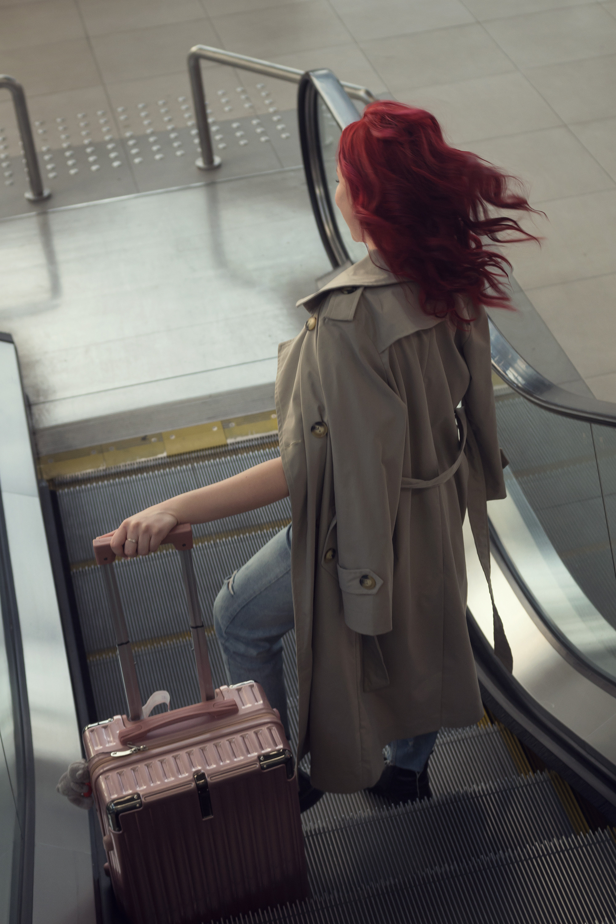 Девушка с чемоданом на эскалаторе. Рыжие волосы развиваются. Вид со спины.