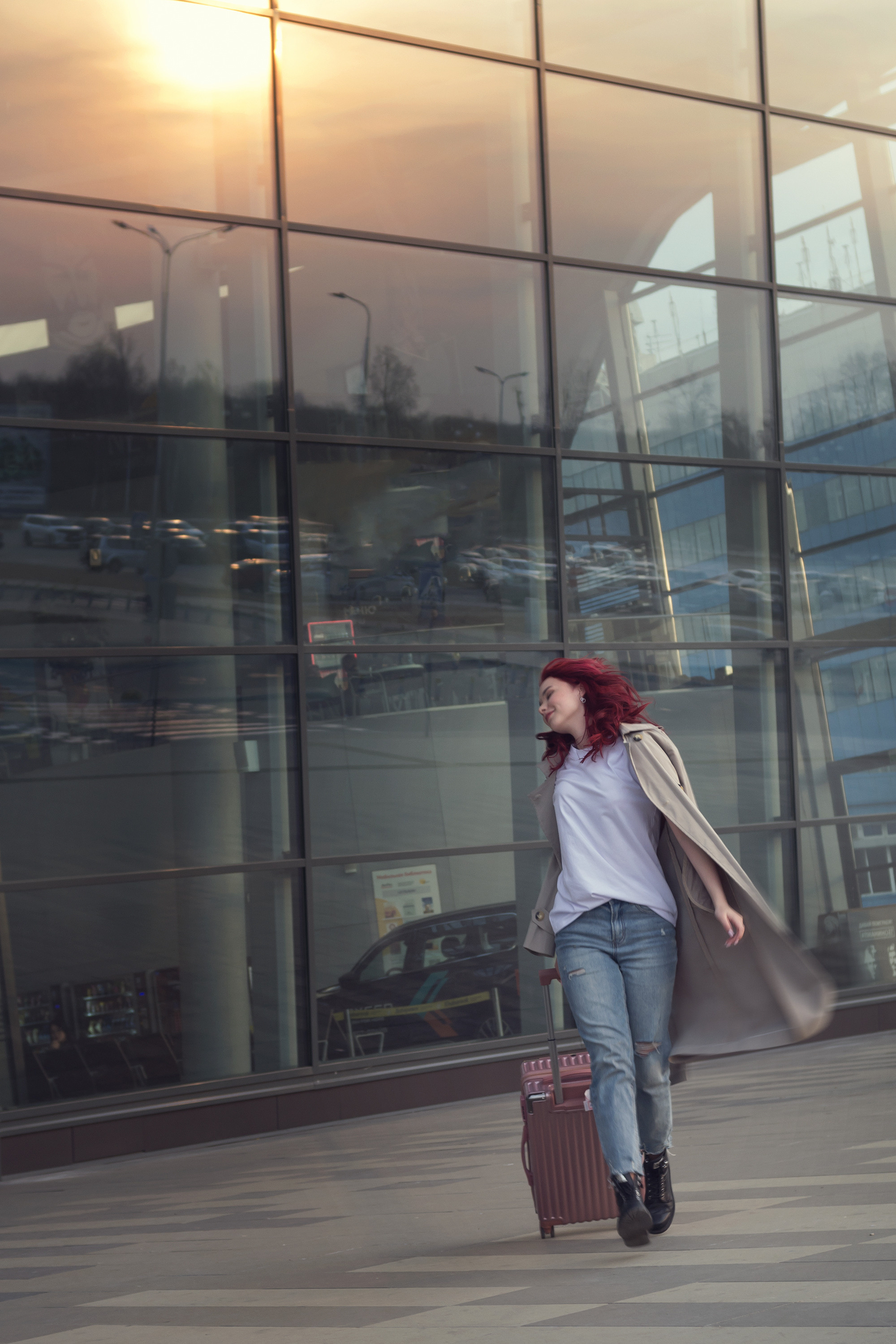 Девушка с чемоданом на фоне аэропорта. В стеклянной стене отражается закат. Одежда кэжуал, джинсы и тренч.