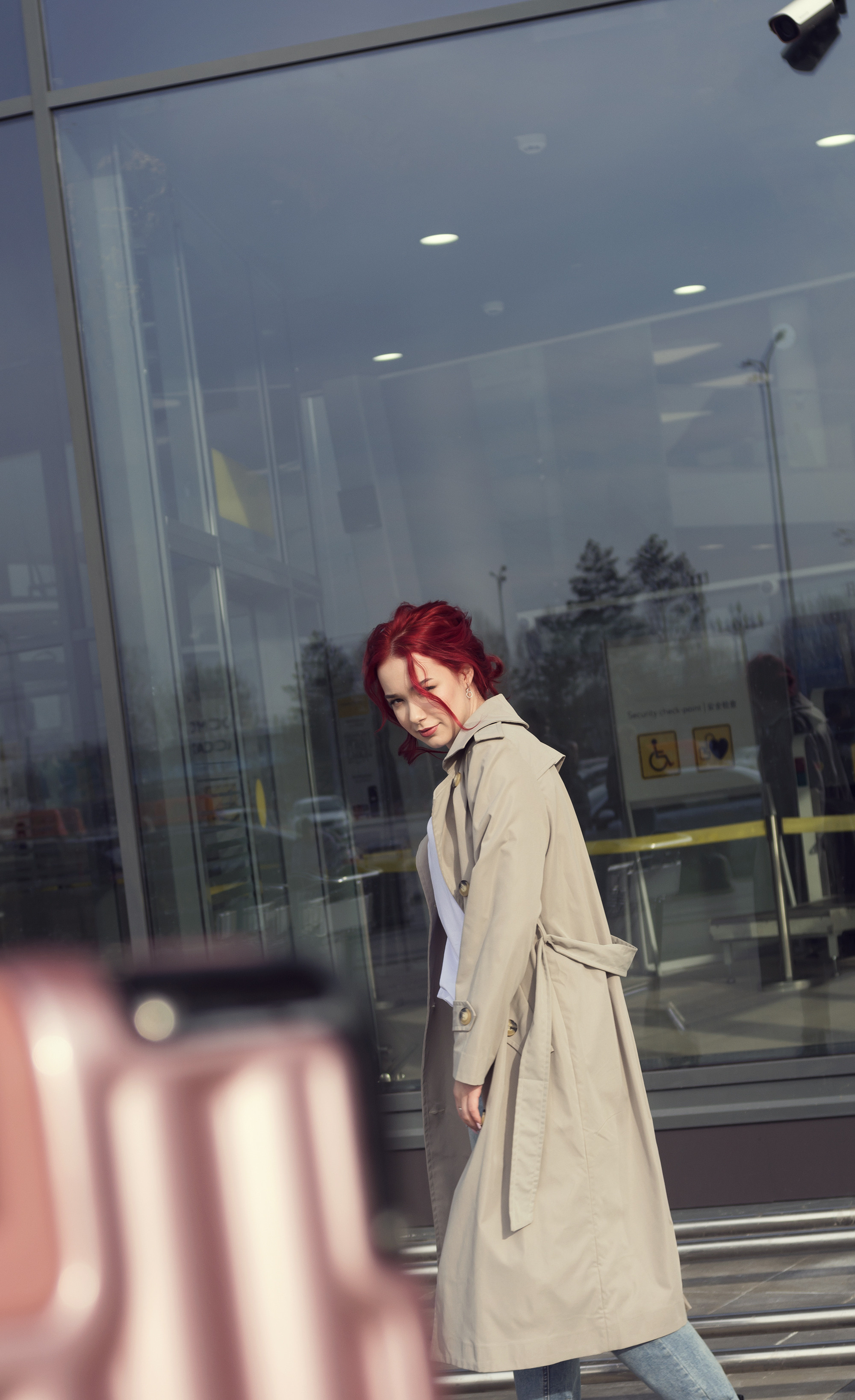 Рыжая девушка в плаще, на фоне стеклянной стены.
